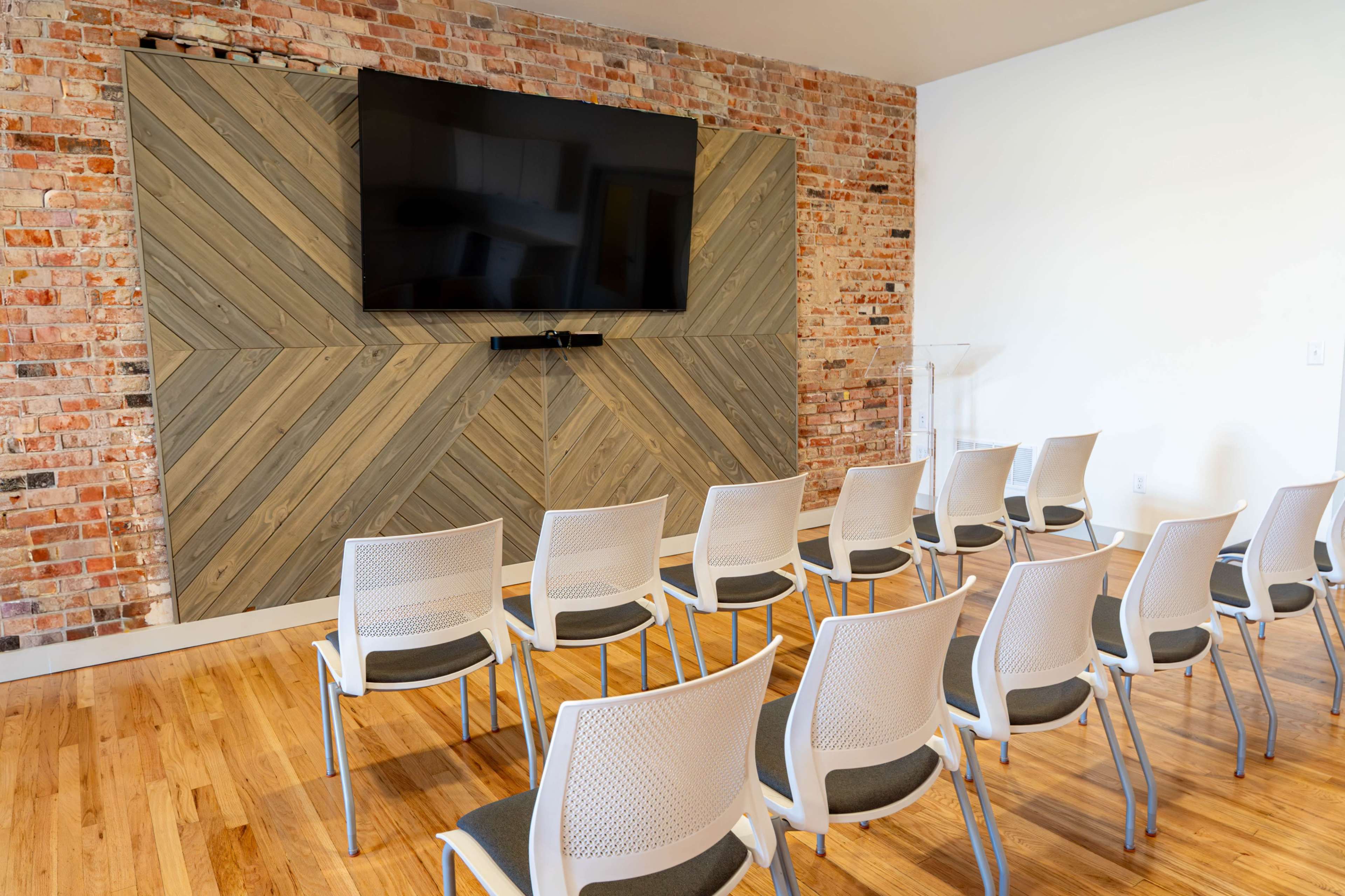 The image shows a meeting room with a large television mounted on a wooden-paneled wall, facing rows of white chairs on a hardwood floor.
