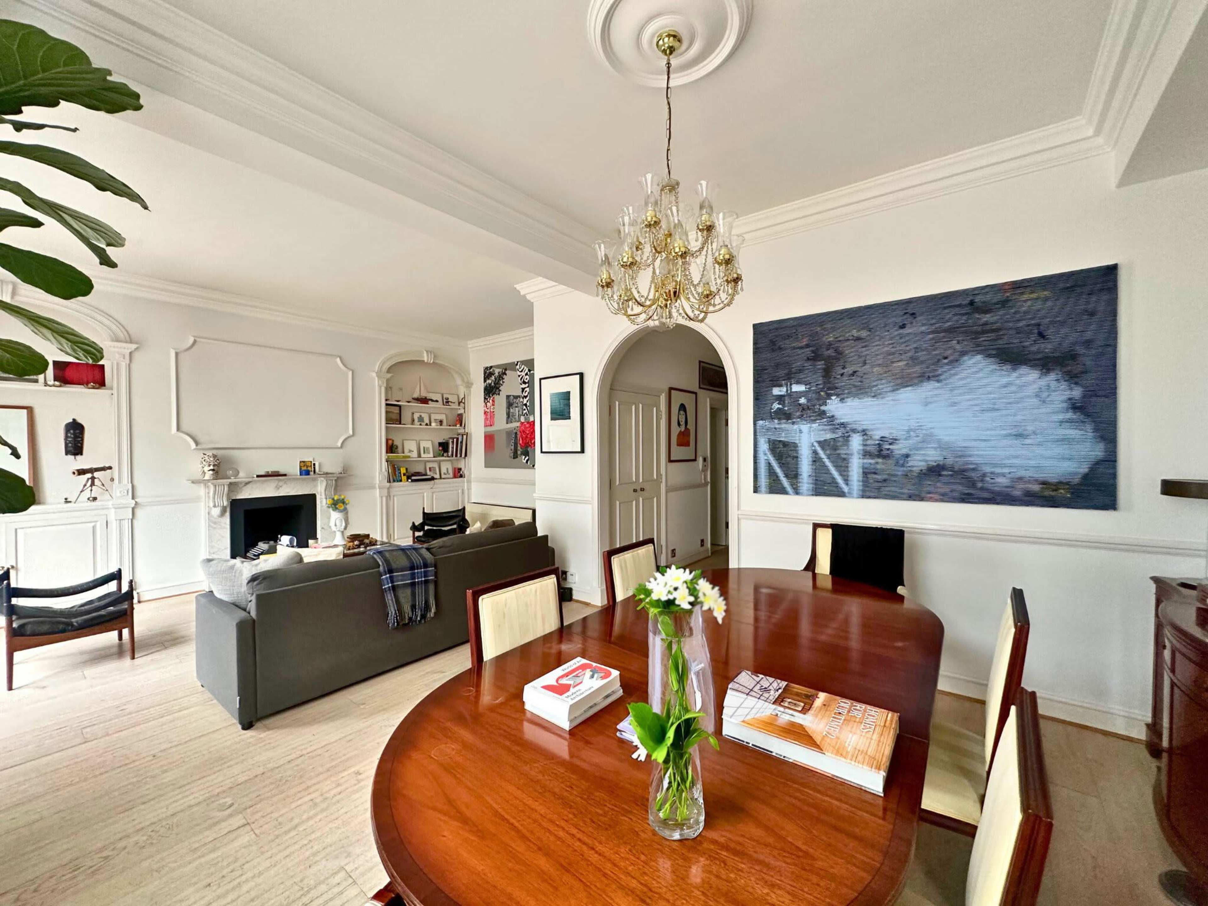 A spacious dining area features a wooden table with flower arrangements, a chandelier overhead, and a living space with a sofa and artwork on the walls.