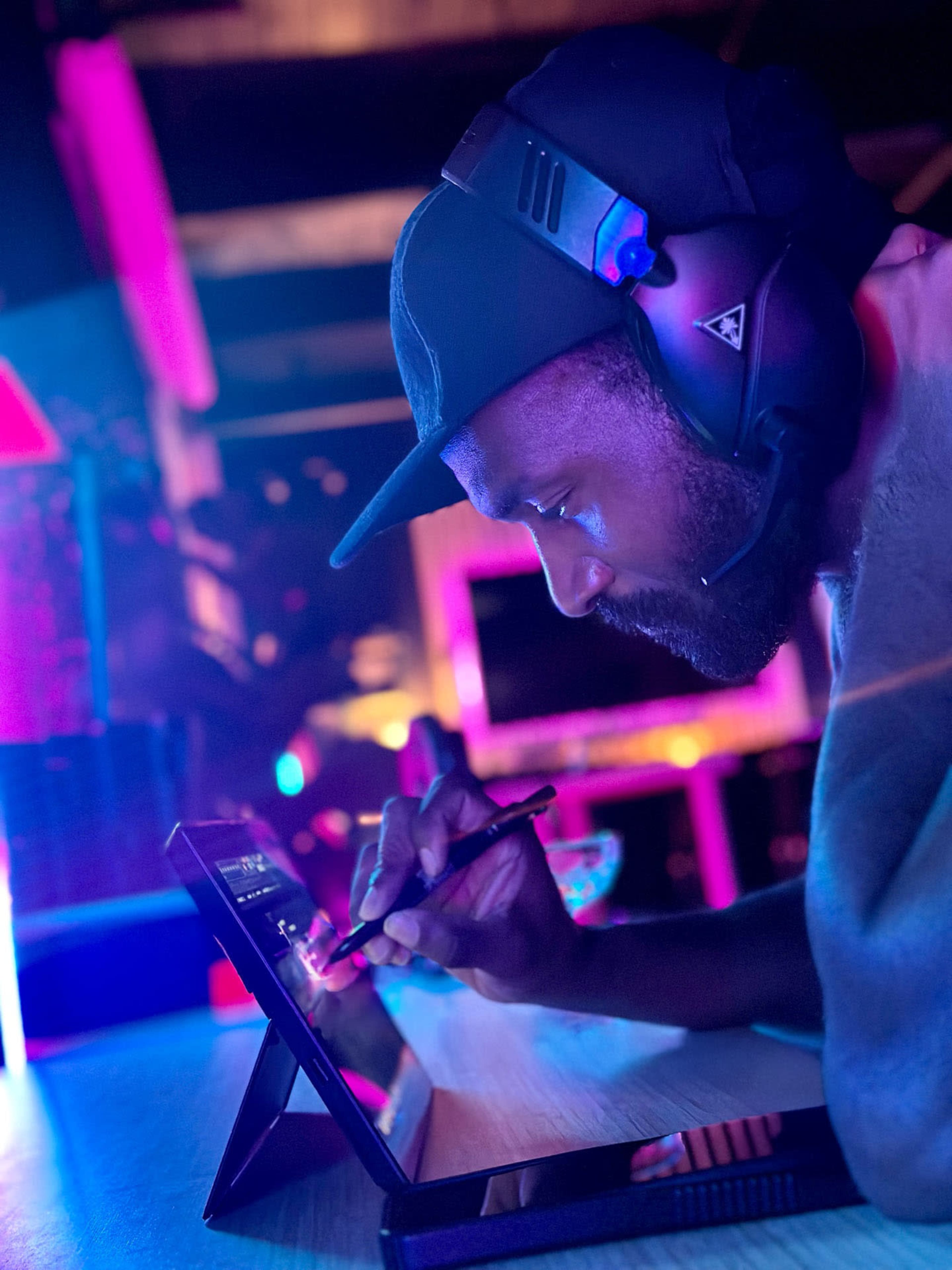 A man wearing a headset is focused on drawing on a tablet in a dimly lit room with colorful lighting.