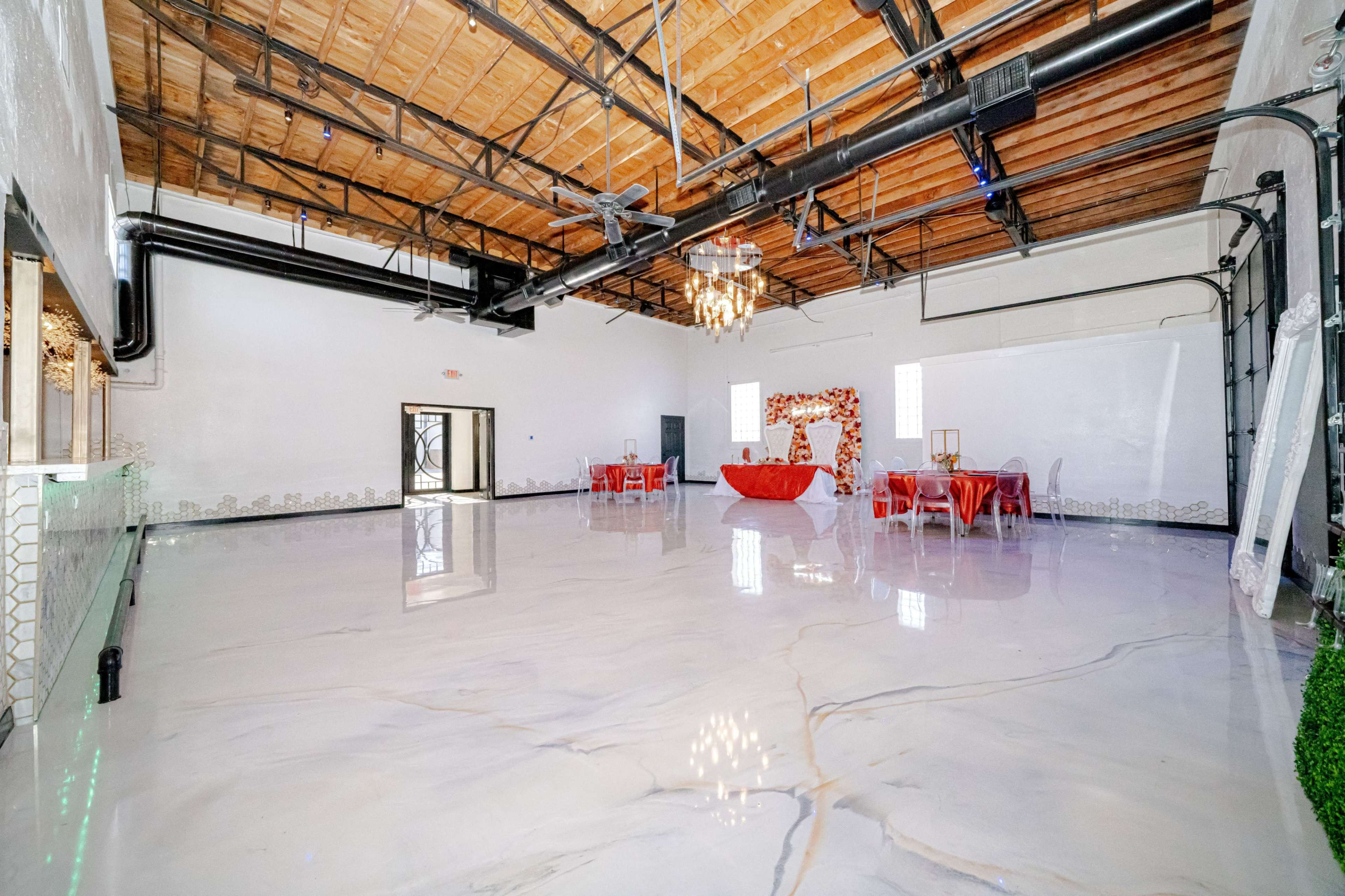 The image shows a spacious, modern event venue with polished floors, exposed wooden beams, and a few tables set up in the center.