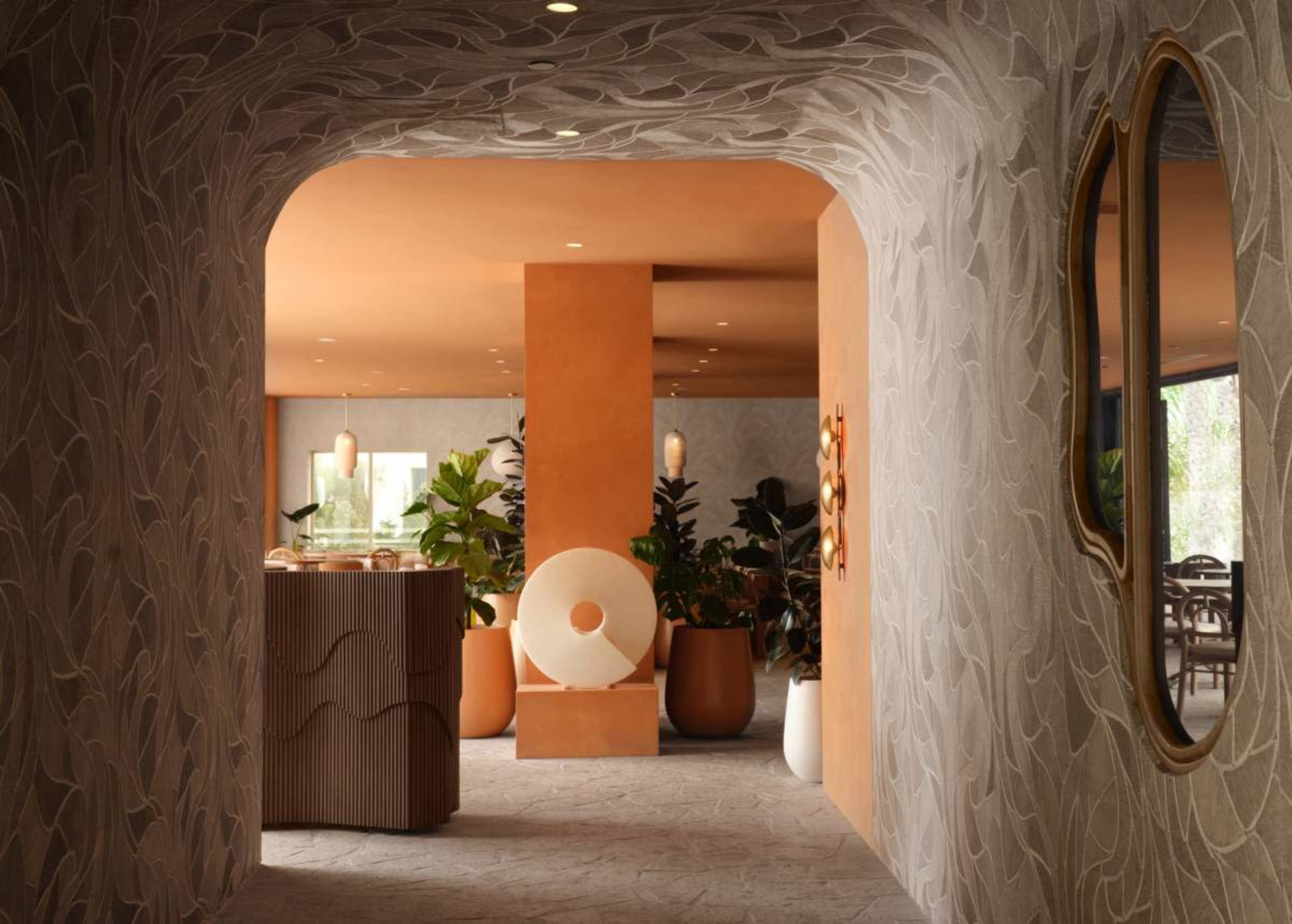 The image features a modern interior space with a decorative hallway, showcasing textured walls, a circular sculpture, potted plants, and warm orange accents.