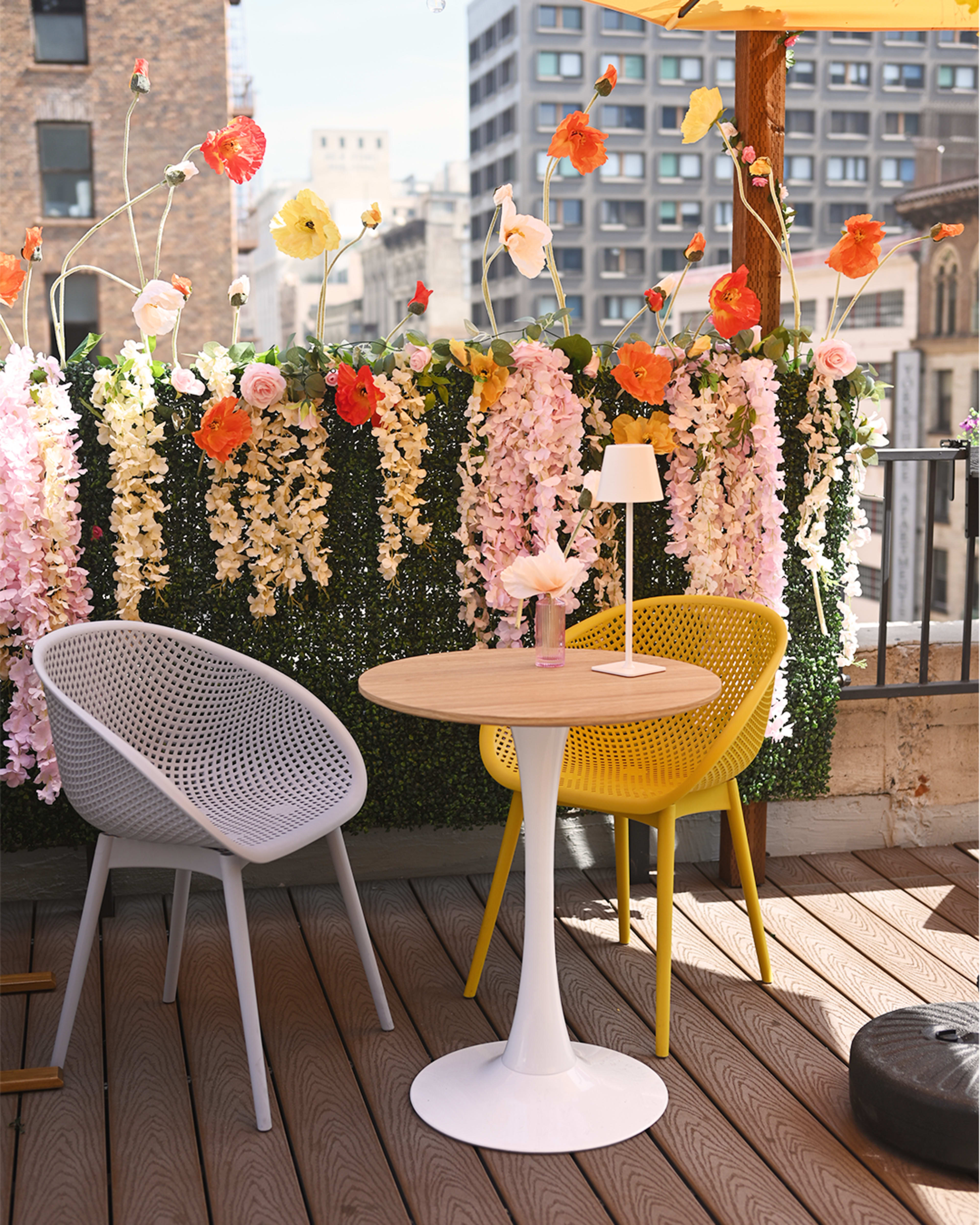The image shows a small outdoor seating area with a white table and two chairs—one gray and one yellow—set against a backdrop of colorful artificial flowers.