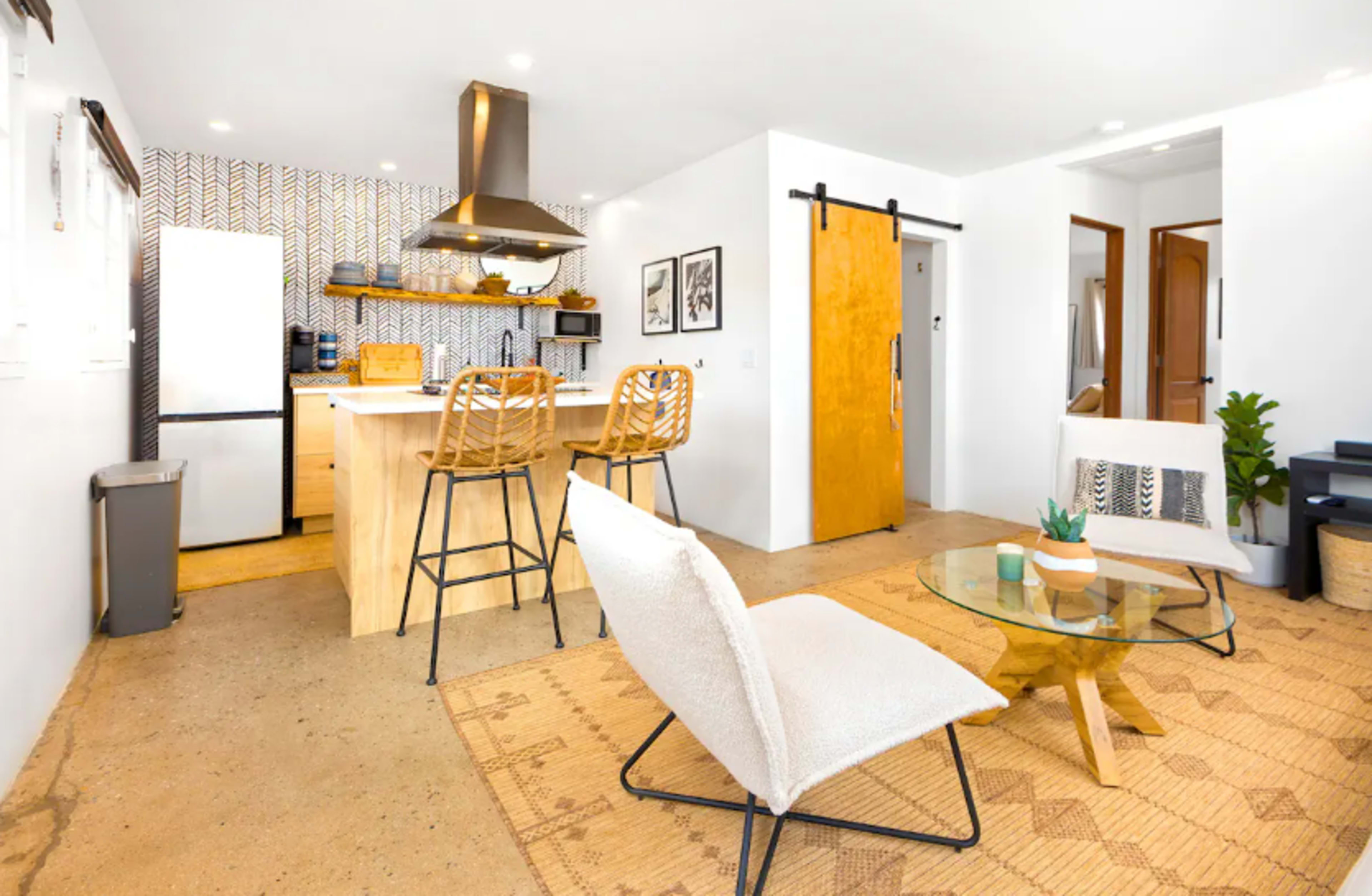 The image shows a modern, open-concept living area featuring a kitchen with wooden cabinetry, a dining table, and minimalist seating arrangements.