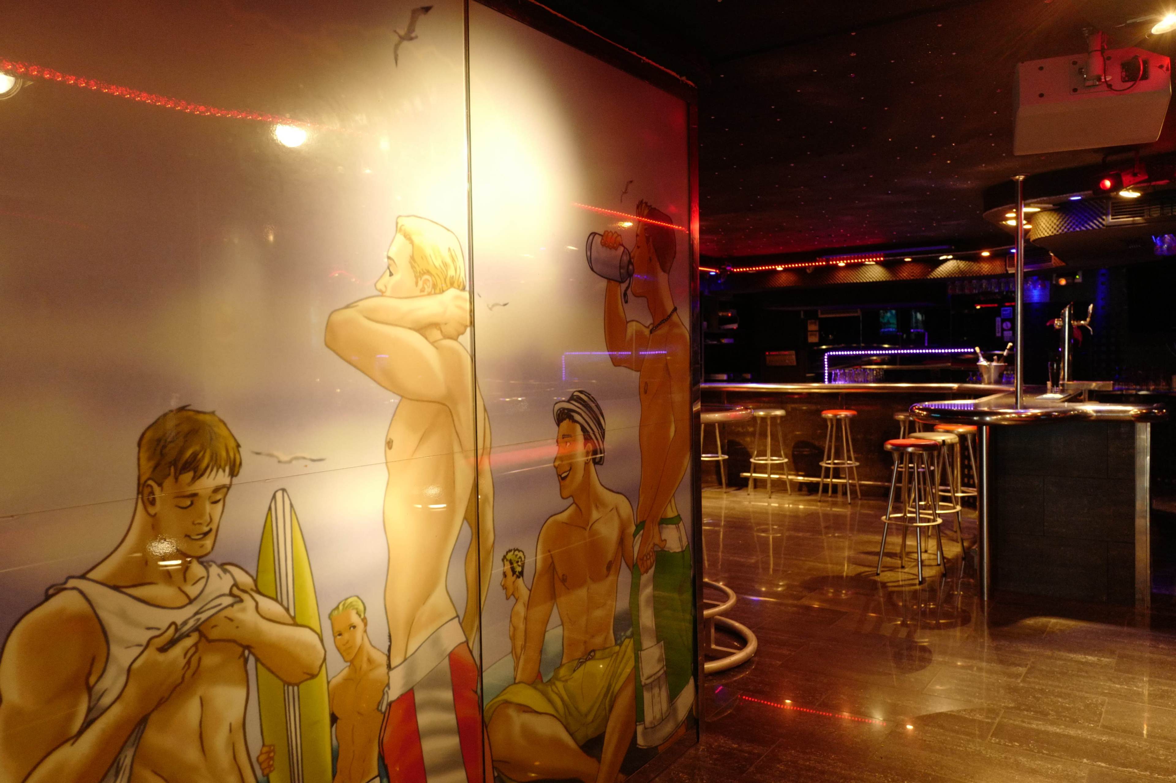 A bar interior with a mural depicting several beach-goers on one wall and bar stools lined up at the counter.