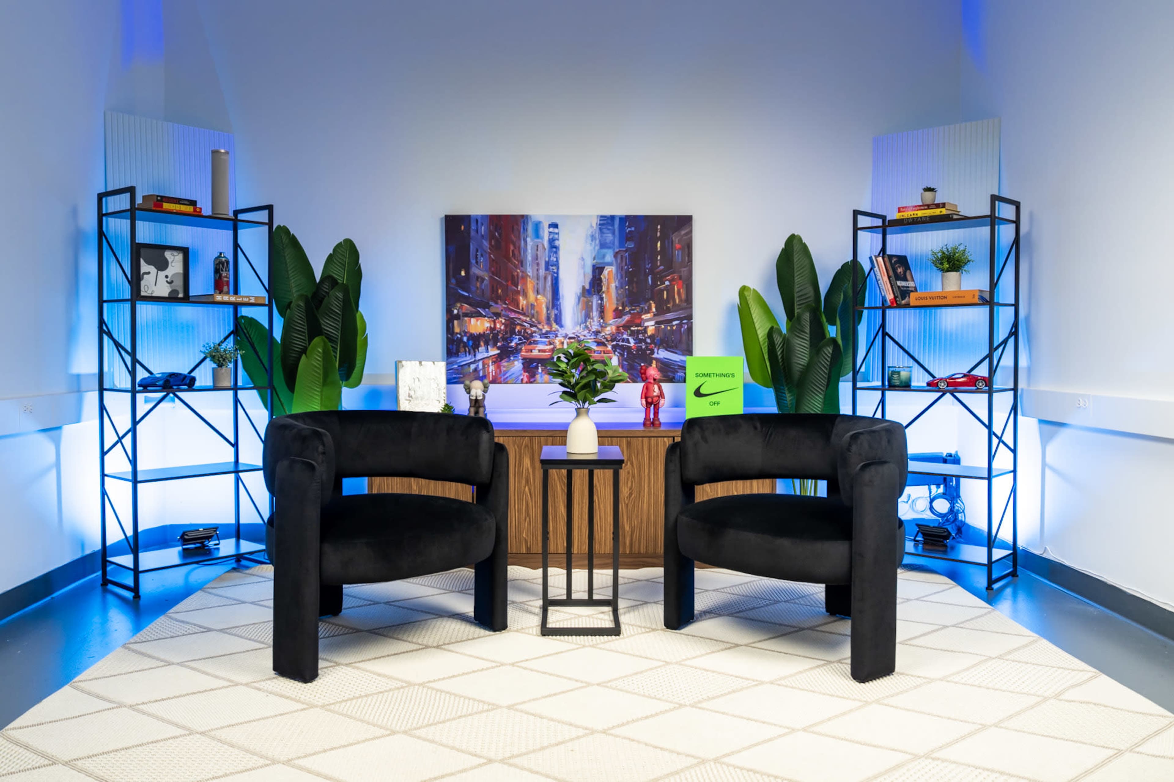 The image features a modern, stylish interior setup with two black chairs facing a wooden console table, surrounded by decorative shelves and potted plants.
