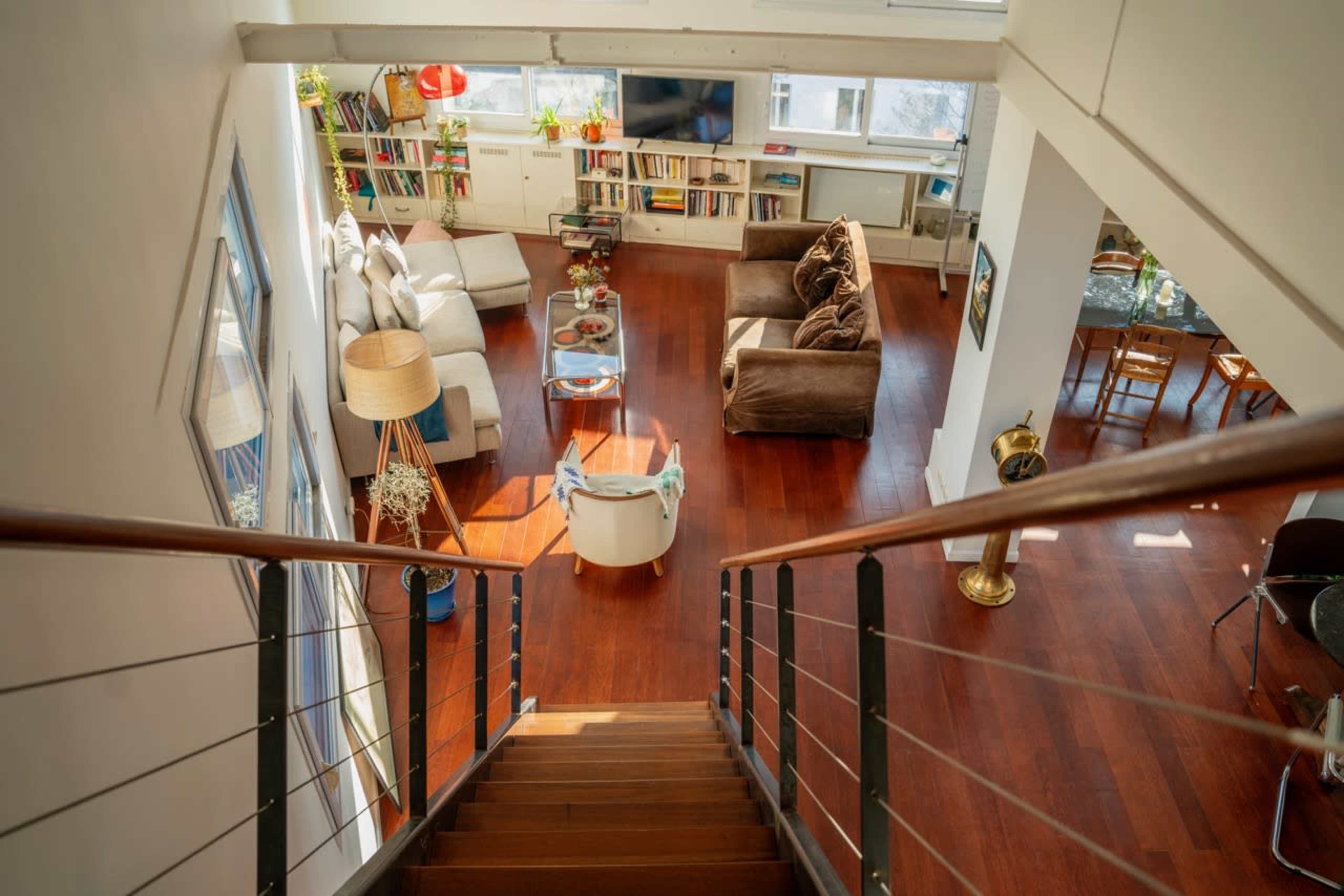 A spacious living area with a brown sofa, a white armchair, shelves filled with books, and wooden flooring, as viewed from the top of a staircase.