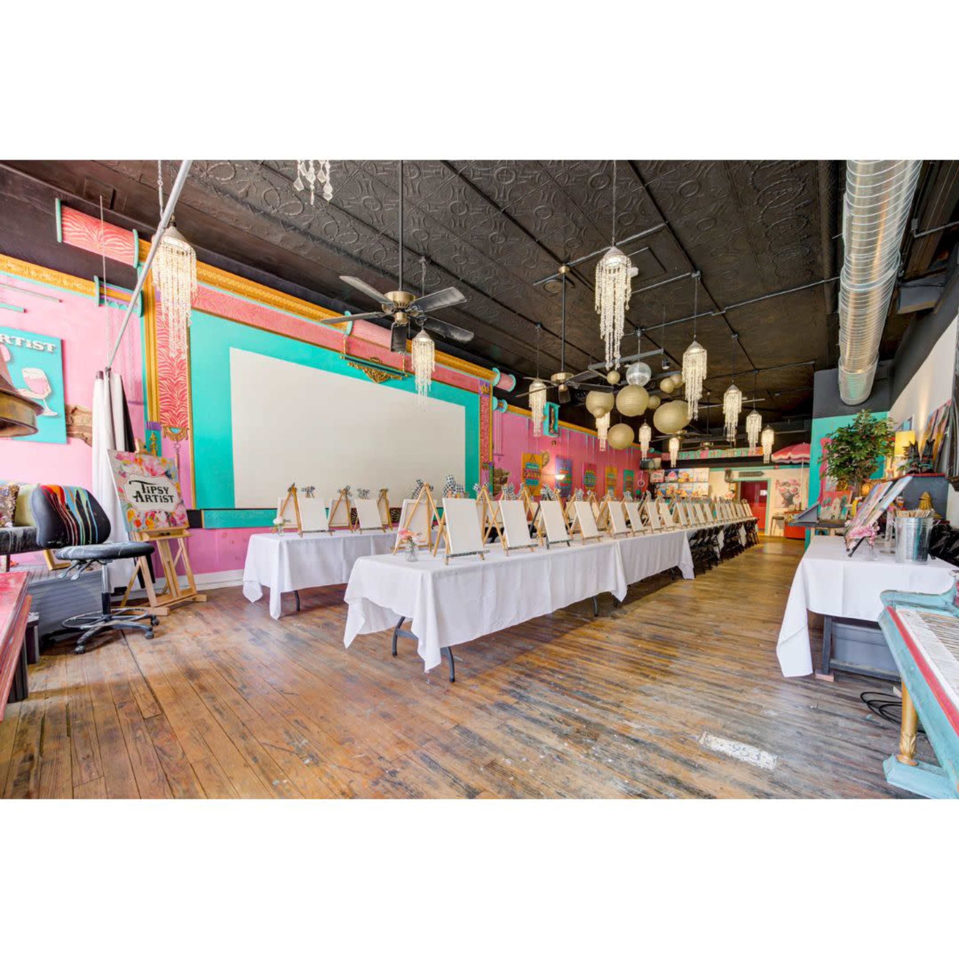 The image shows an art studio setup with multiple easels and blank canvases arranged along a row of tables in a colorful, well-lit space.