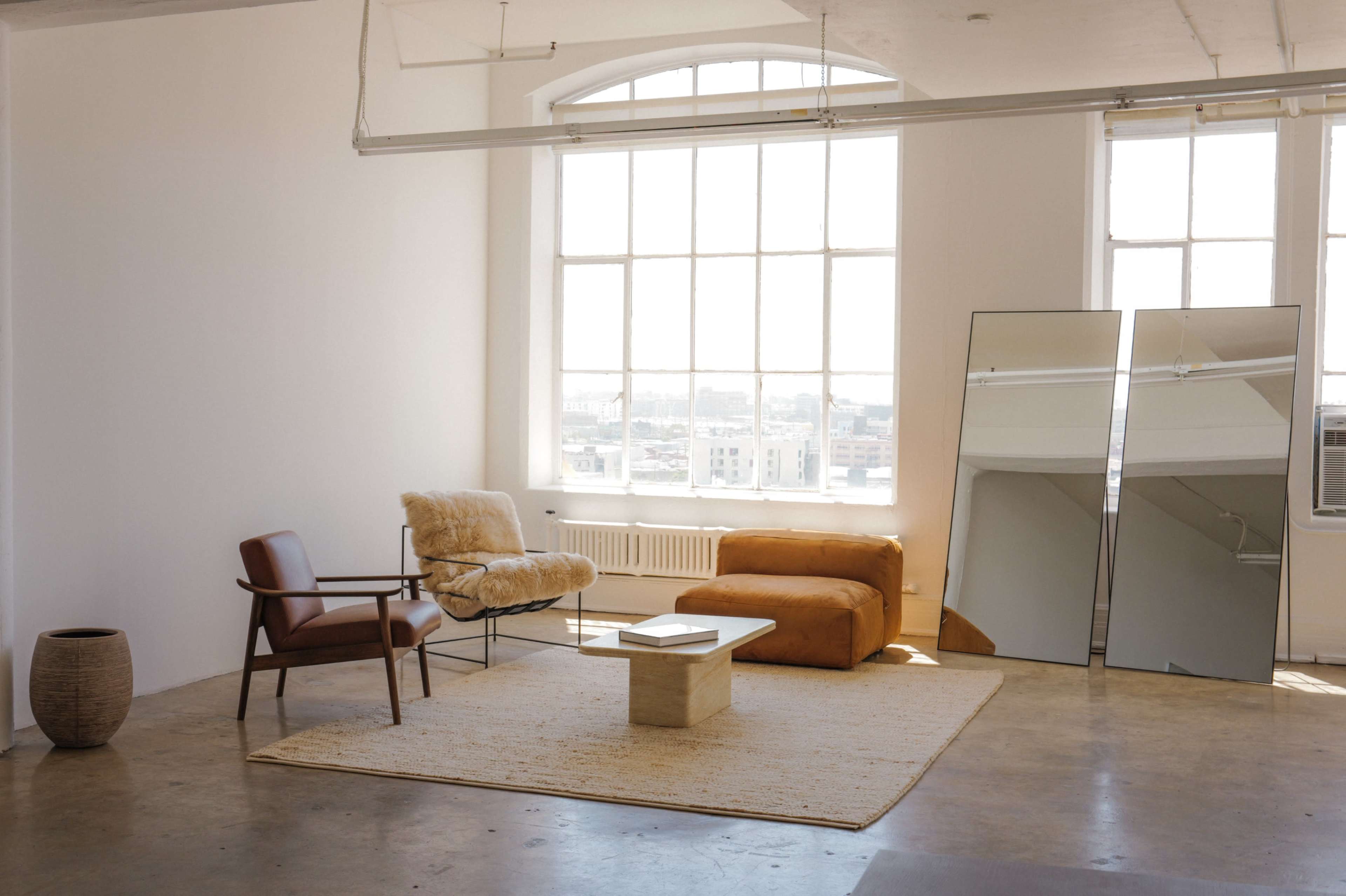The image shows a spacious, minimalist interior with large windows, a brown leather sofa, a cozy chair, a coffee table, and two tall mirrors against a wall.