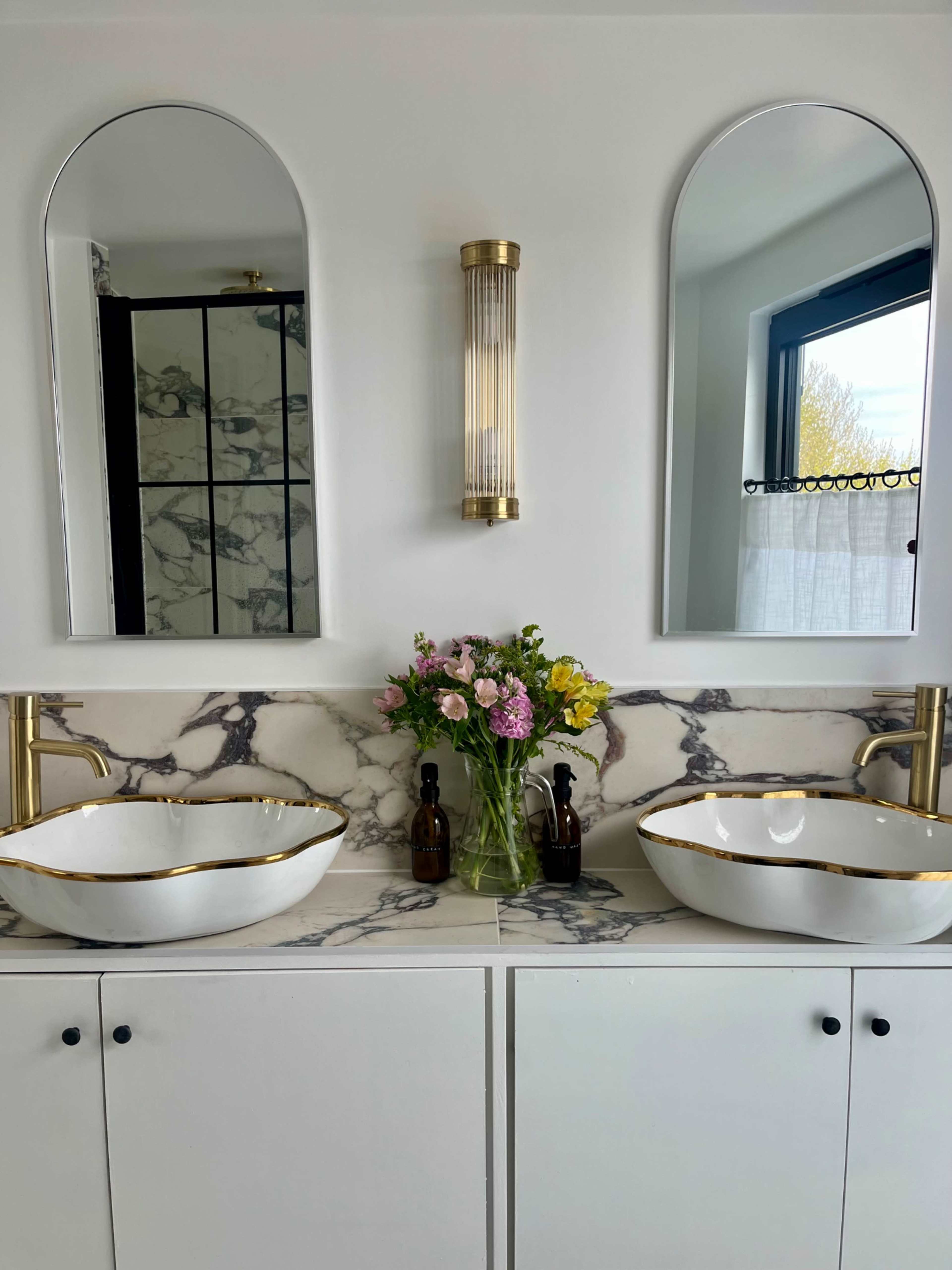 The image shows a modern bathroom with two round mirrors above separate white and gold-accented sinks, a floral arrangement in the center, and a stylish light fixture on the wall.
