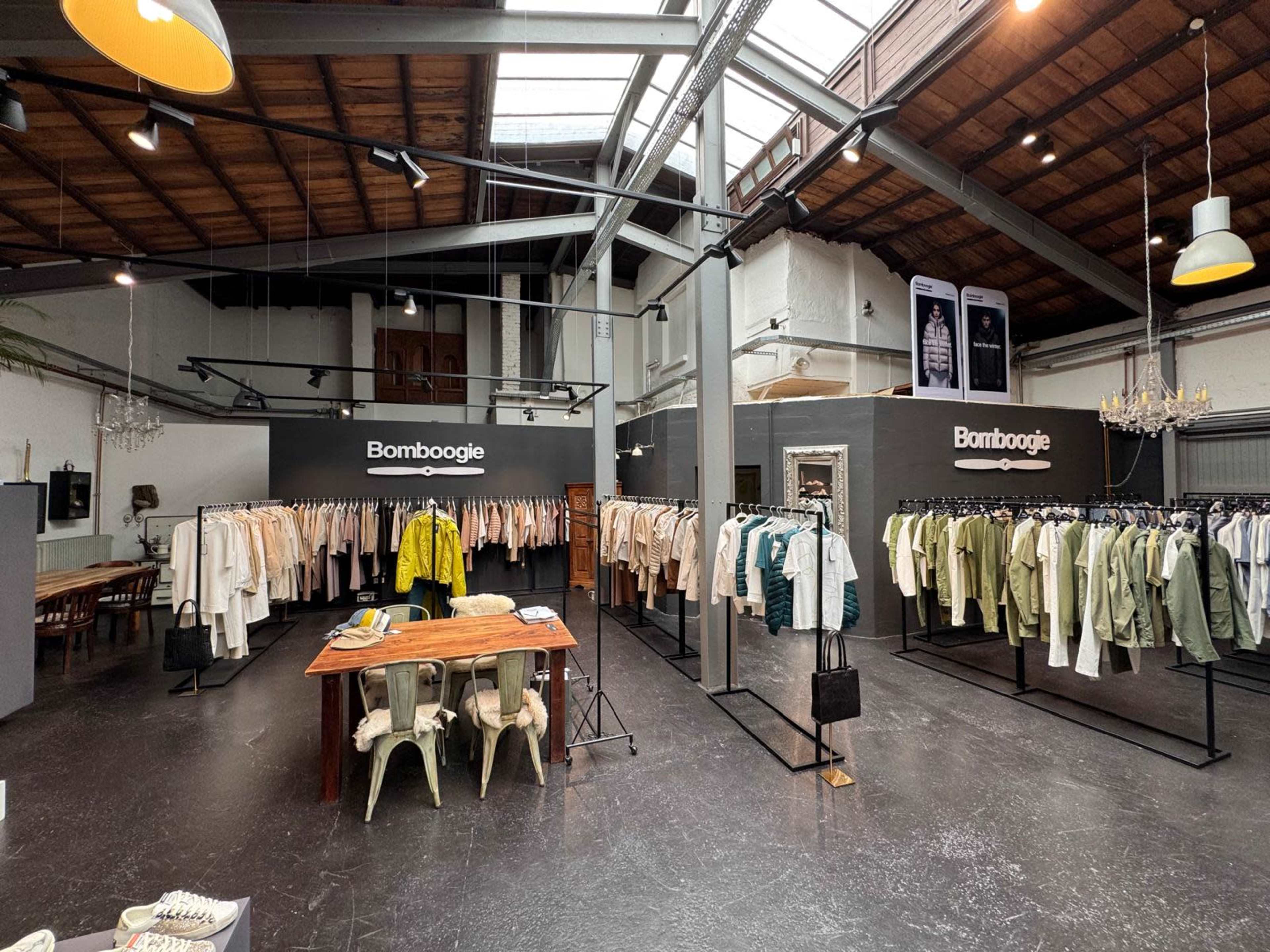 The image shows a clothing store interior with racks of garments arranged neatly, featuring a central wooden table and a high ceiling with industrial-style lighting.