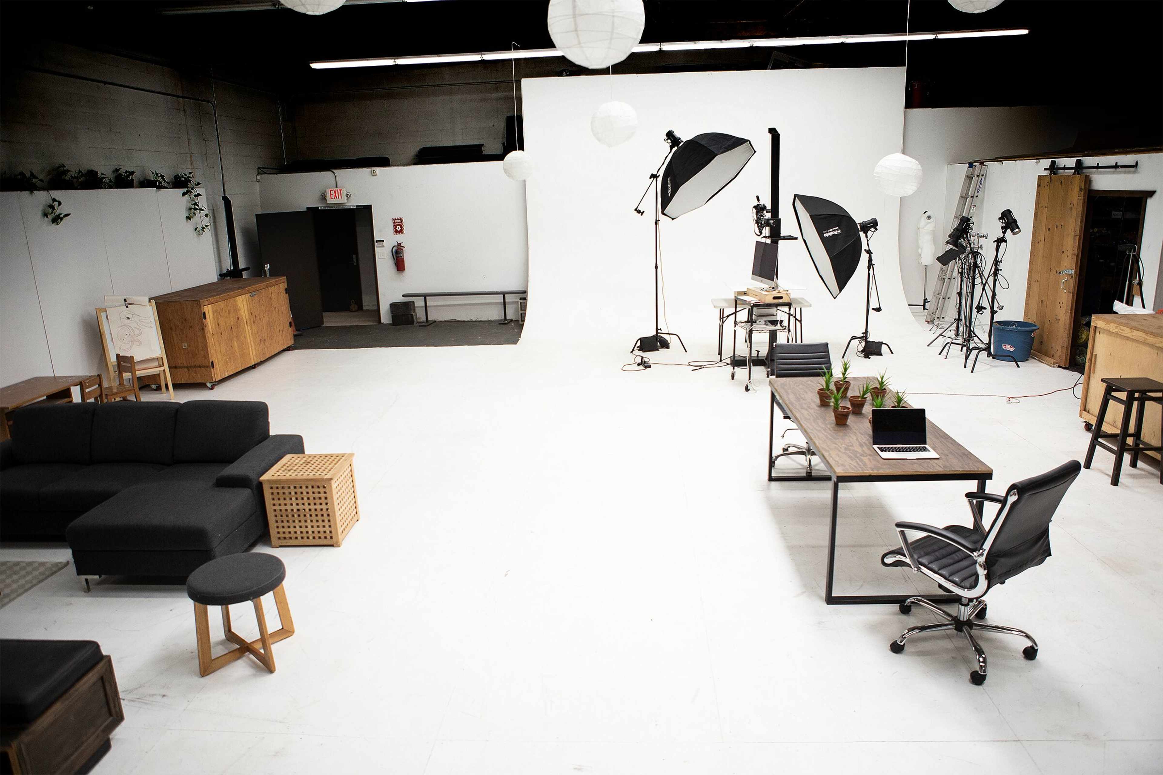 A spacious, well-lit studio with a plain backdrop, multiple photography lights, and a variety of furniture arranged for a photo shoot.