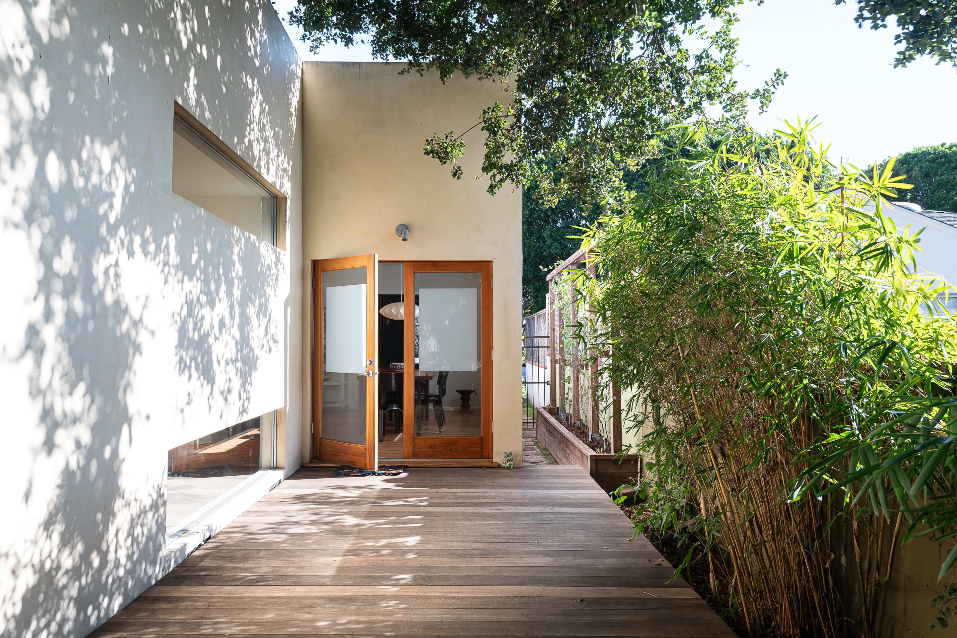 A wooden door opens onto a sunlit deck surrounded by greenery and a textured wall.