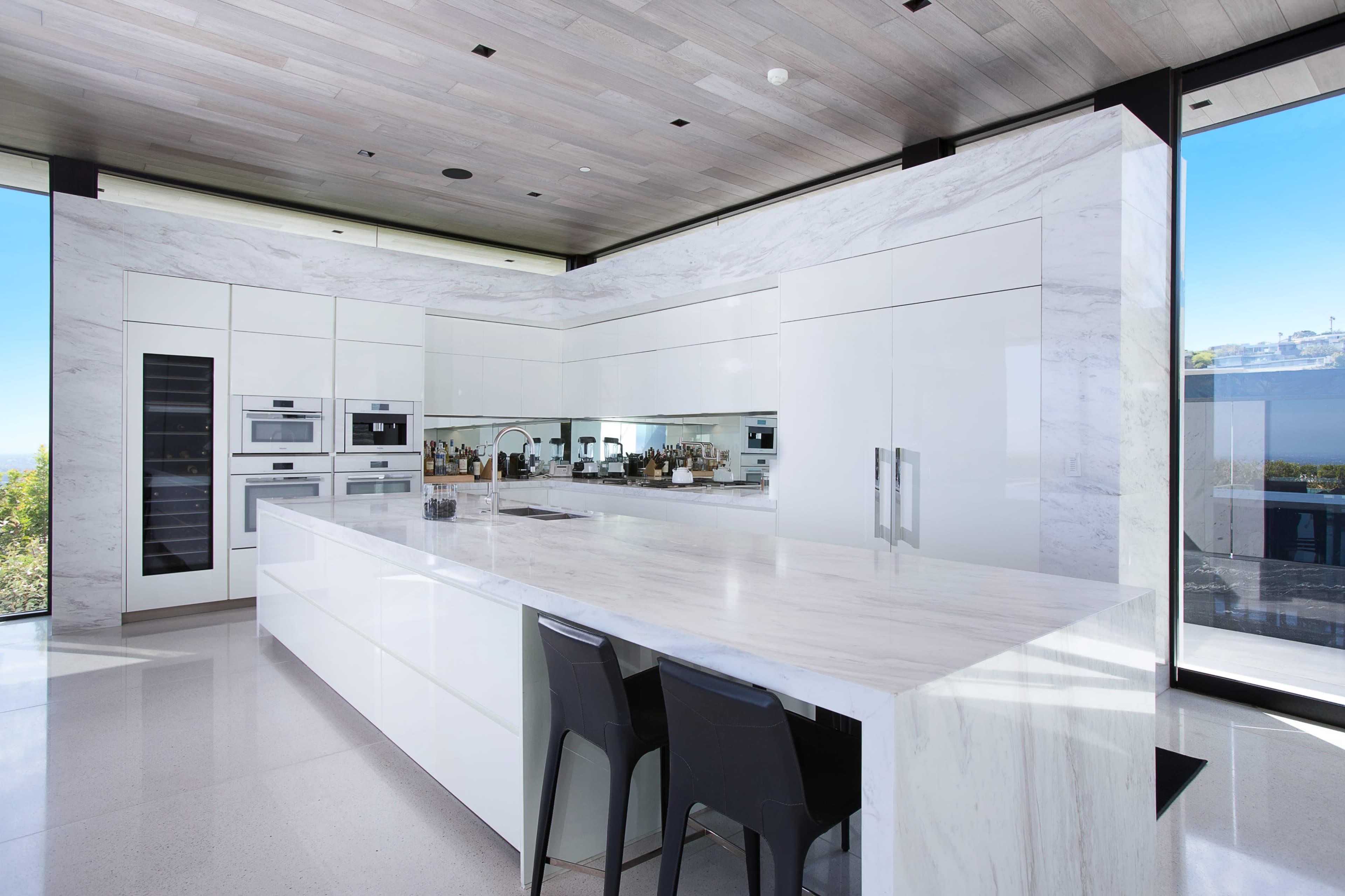 The image shows a modern kitchen featuring a large white marble island, sleek cabinetry, built-in appliances, and floor-to-ceiling windows.