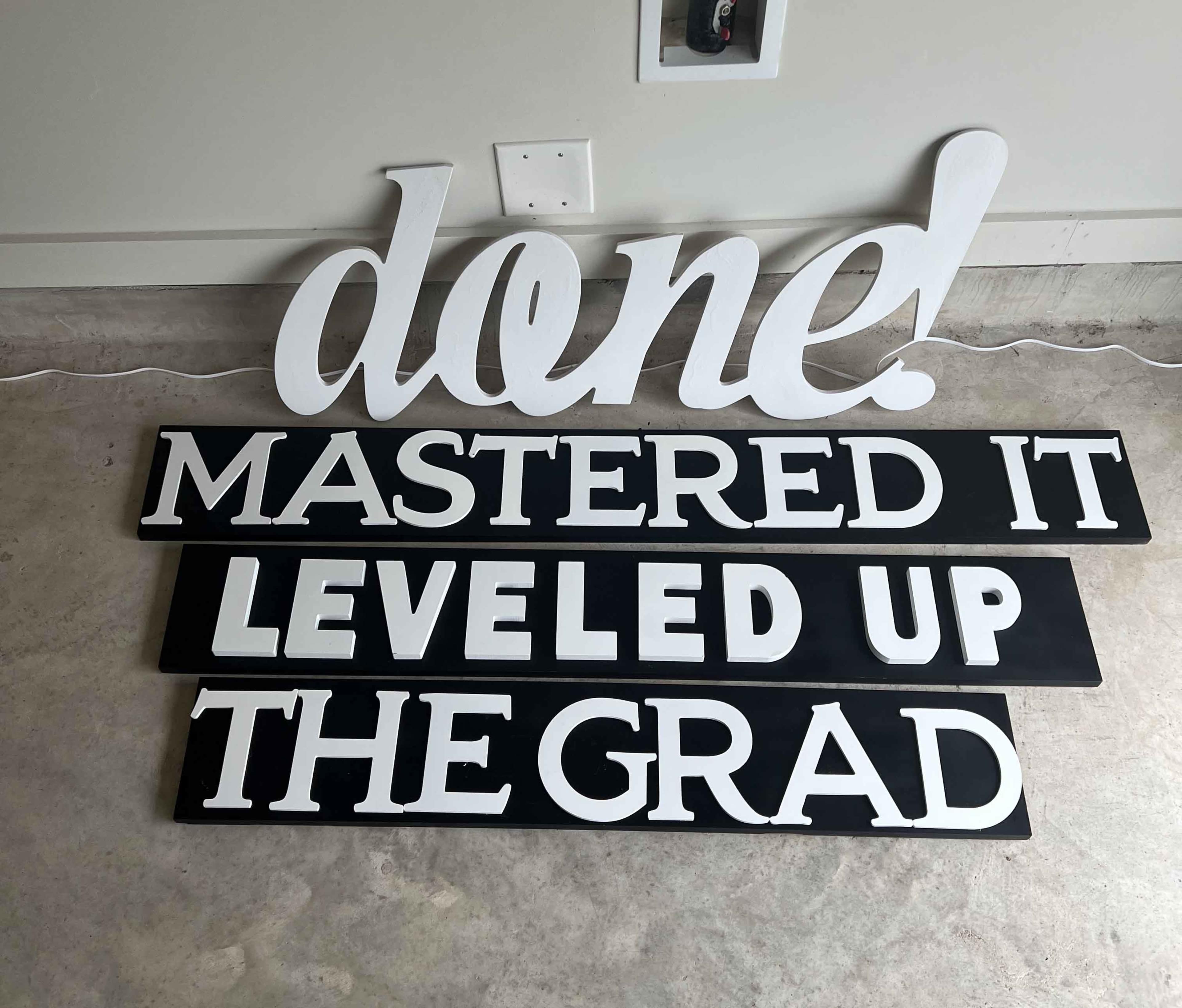 The image shows a group of large, decorative sign letters that spell out phrases related to graduation, with the words "done," "MASTERED IT," "LEVEL UP," and "THE GRAD."