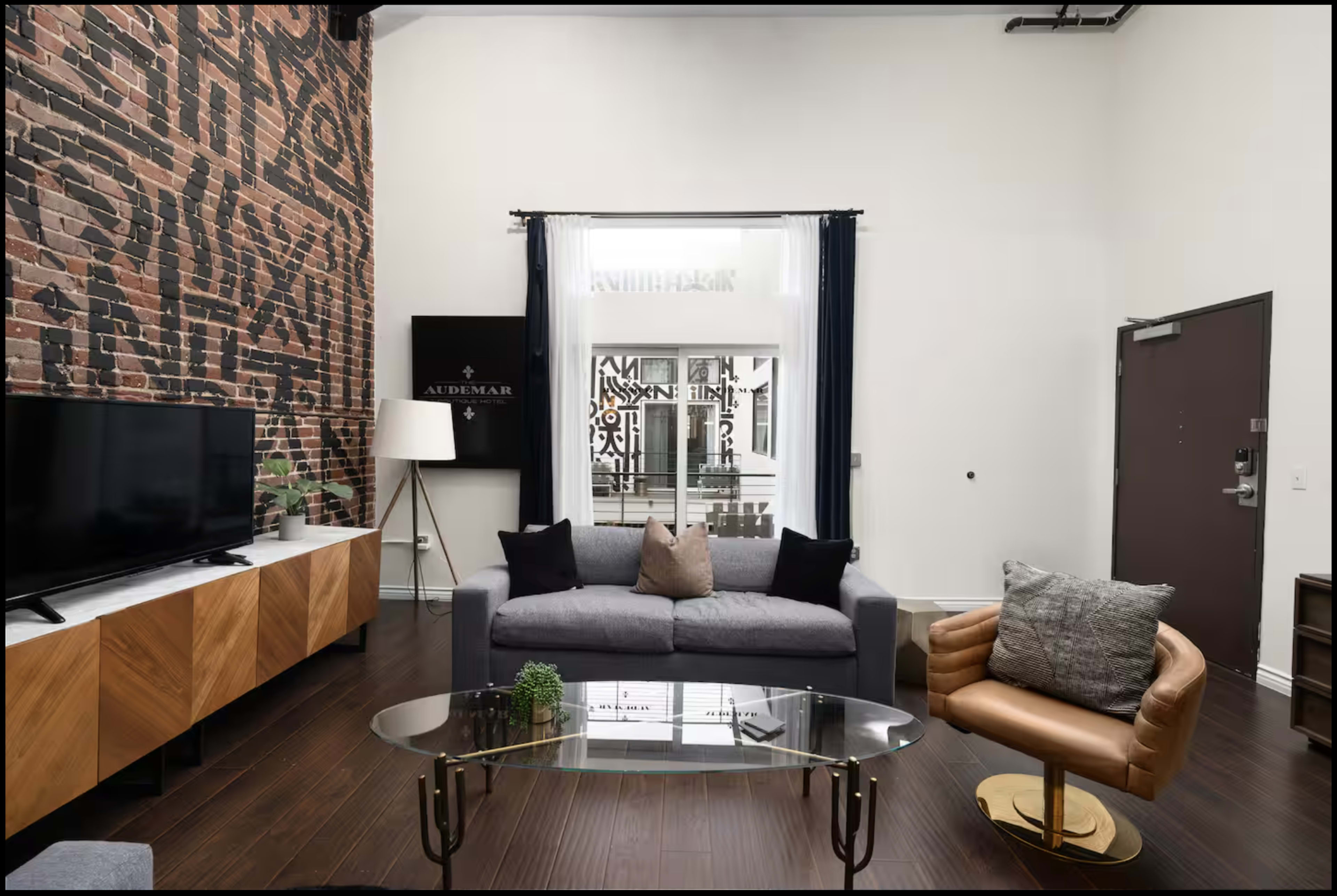 The image shows a living room with a large brick wall, a gray sofa with decorative pillows, a glass coffee table, a TV stand with a television, and a stylish armchair.
