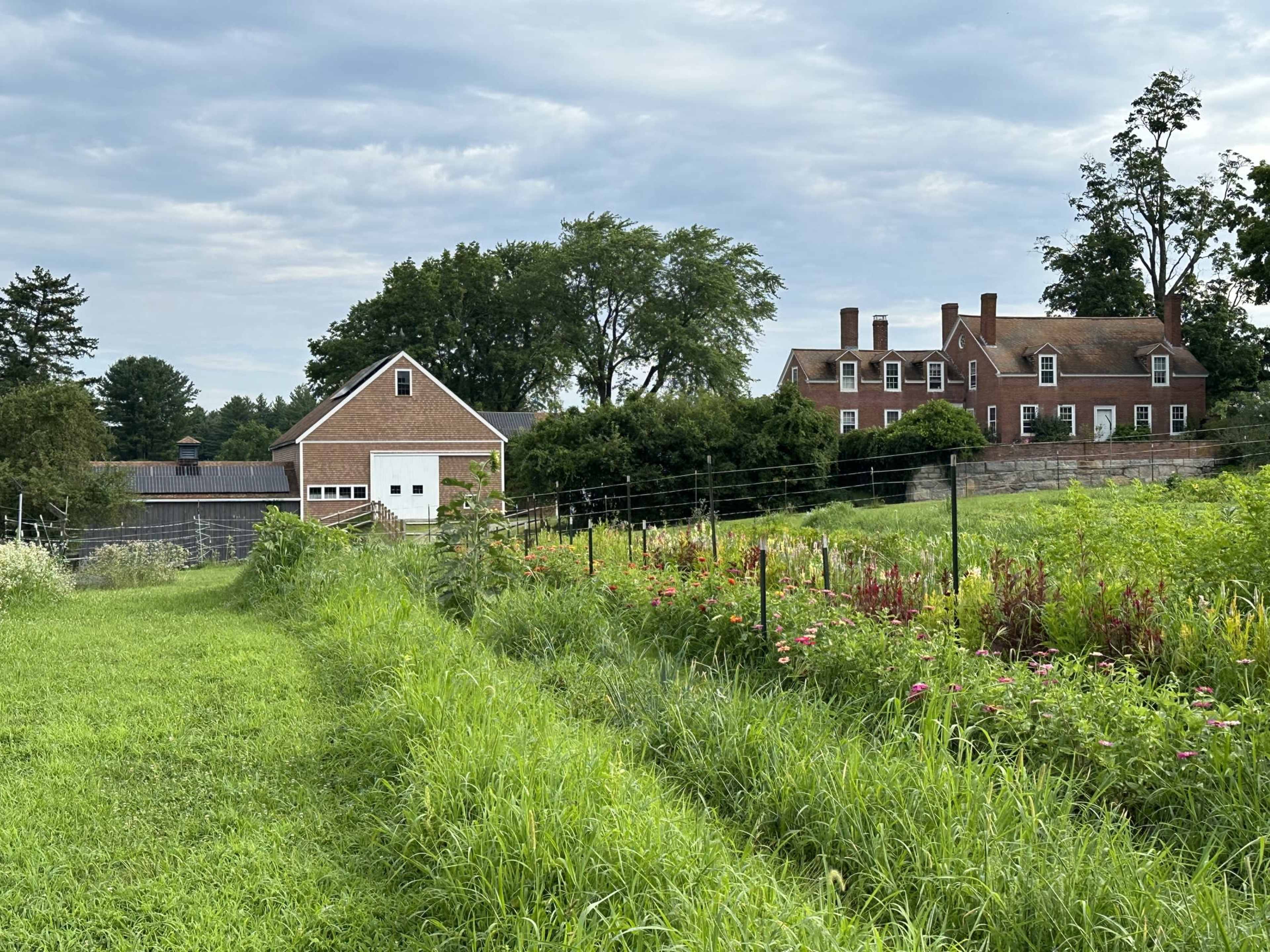 The image shows a green field leading to a large brick house and a nearby barn, with trees in the background and a cloudy sky above.