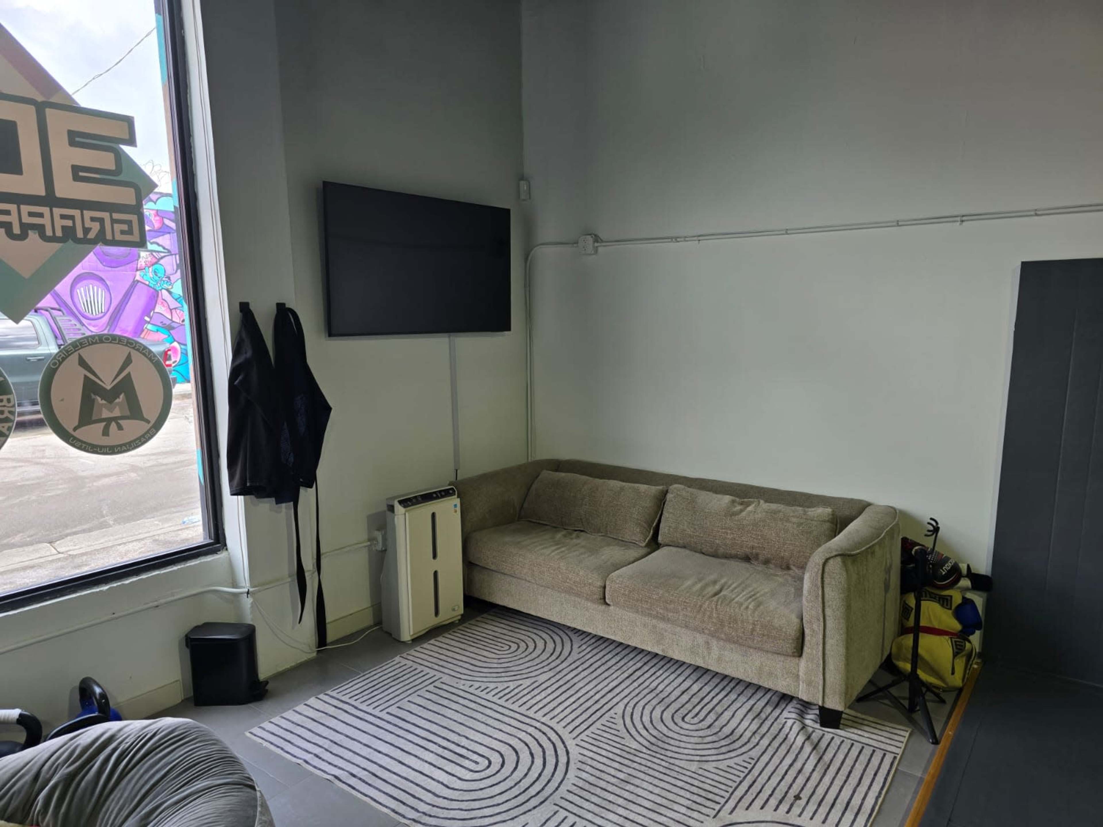 The image shows a small, minimally furnished room featuring a gray couch, a wall-mounted television, and a window displaying colorful artwork.
