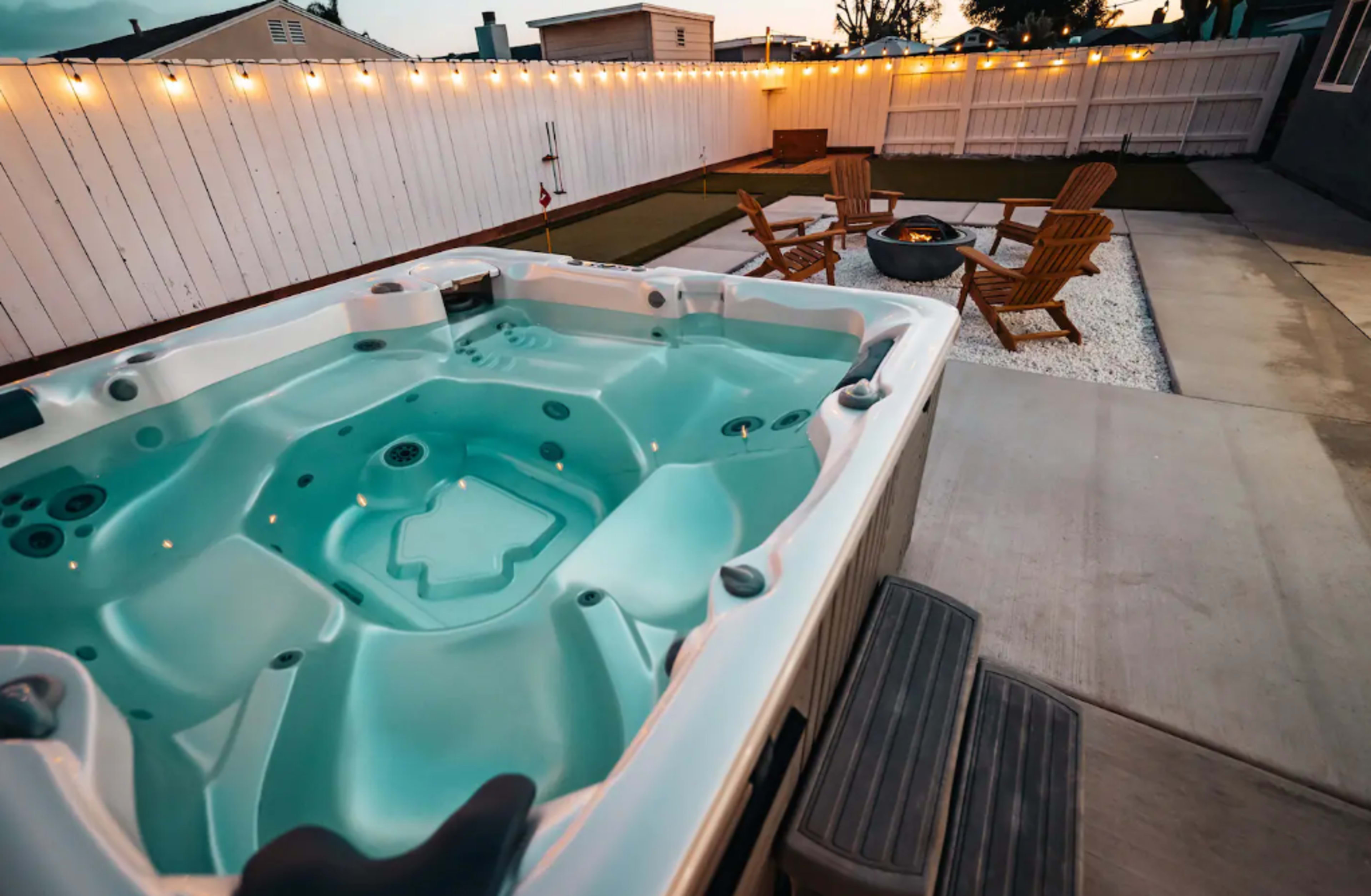 A hot tub is situated next to a fire pit and wooden chairs in a yard surrounded by a white fence, under string lights.