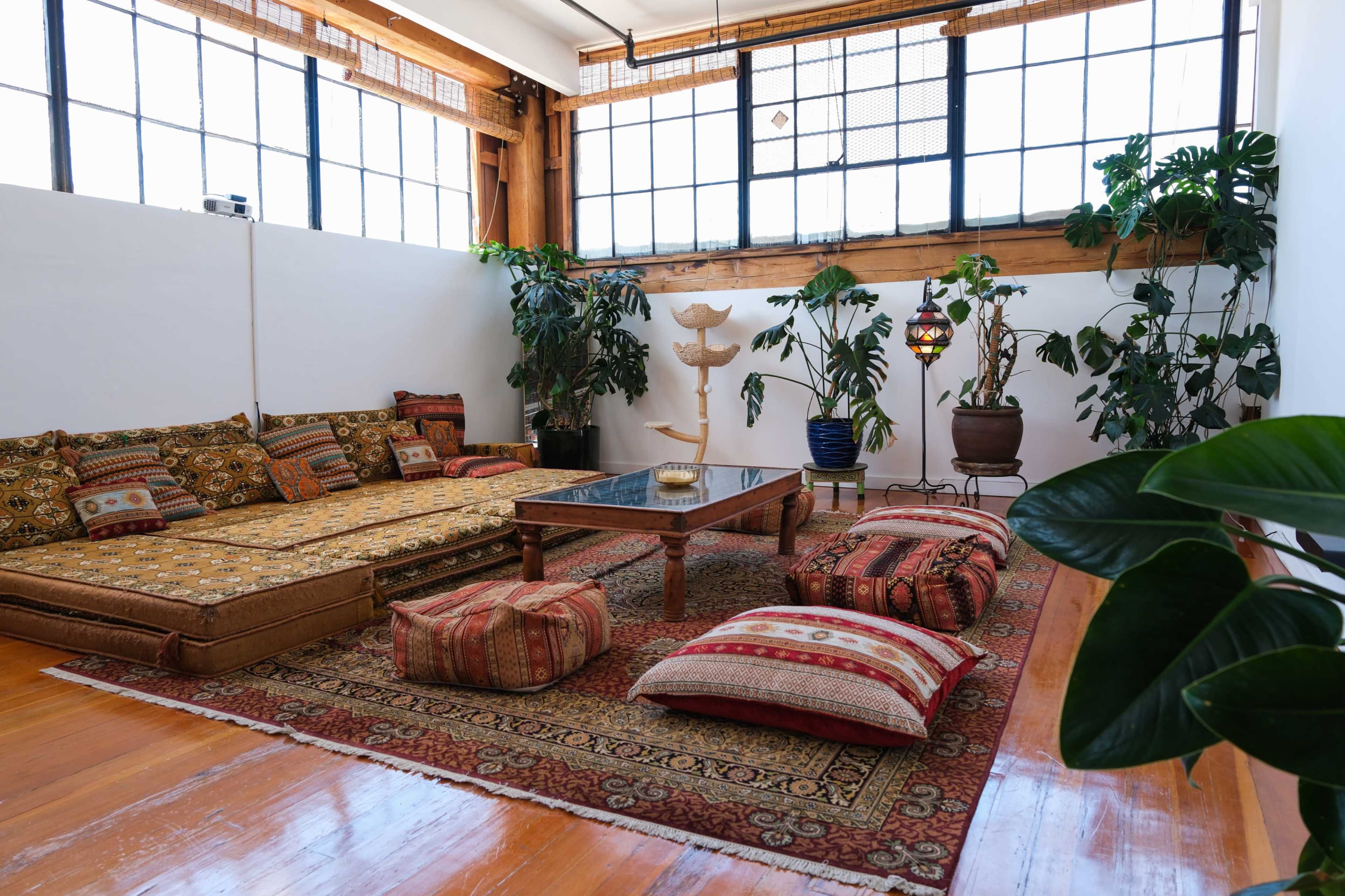 A spacious room features a low seating area with patterned cushions and a central table, surrounded by various plants and illuminated by natural light from large windows.