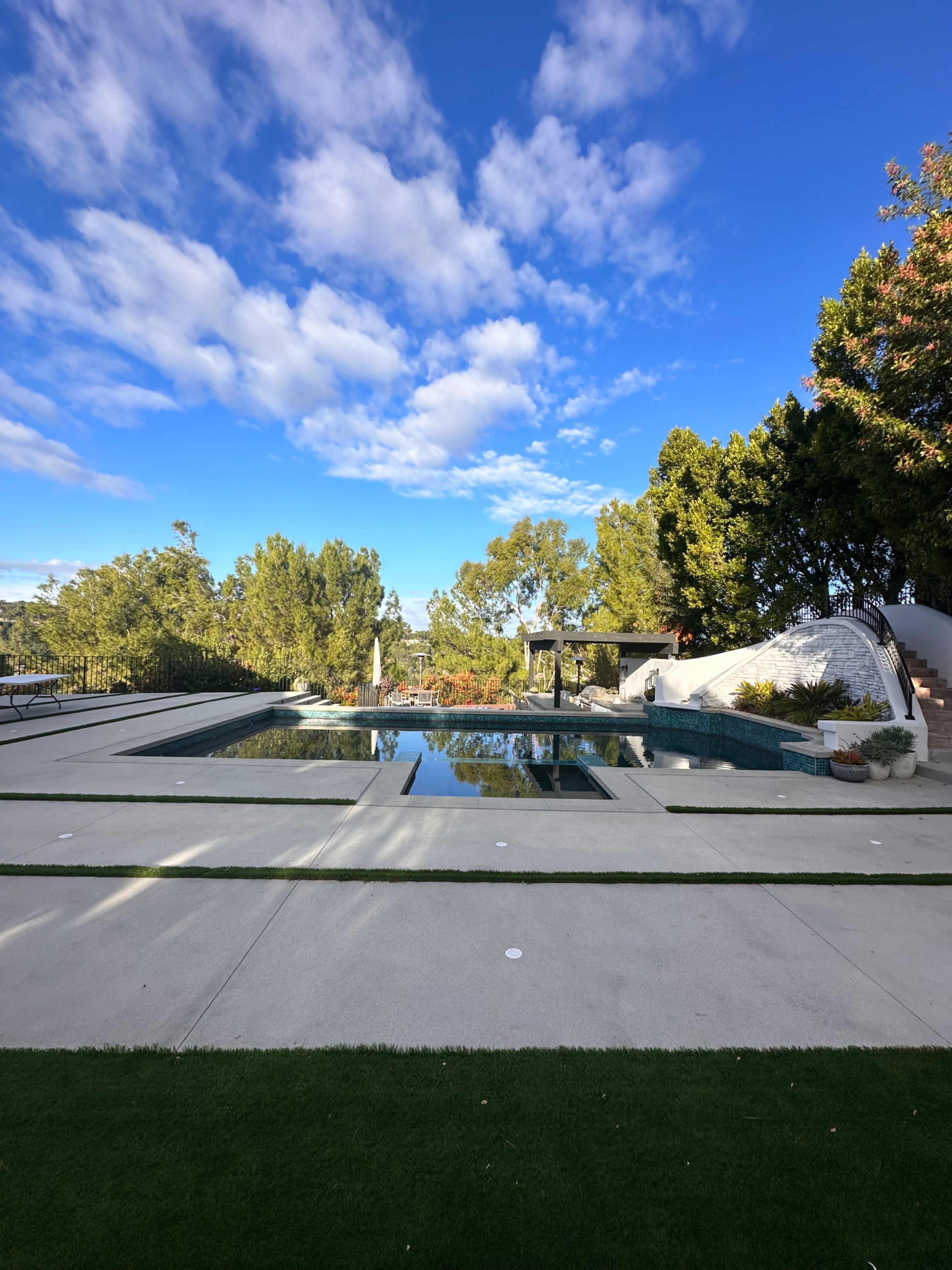 A modern outdoor pool area features a large swimming pool surrounded by concrete decking and landscaped greenery under a blue sky with scattered clouds.