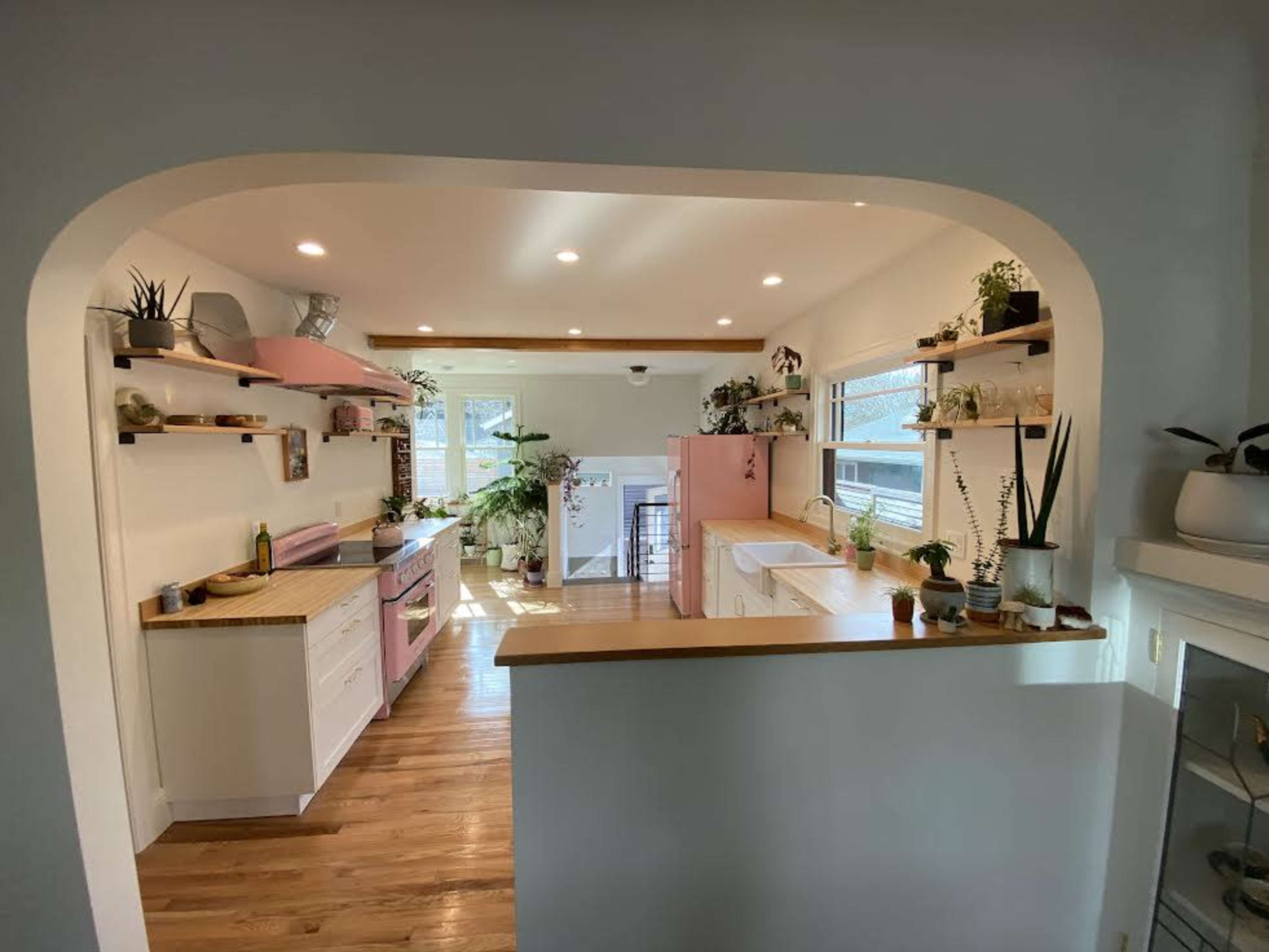 A view of a kitchen featuring pink and white cabinetry, wooden countertops, and ample greenery displayed on shelves.