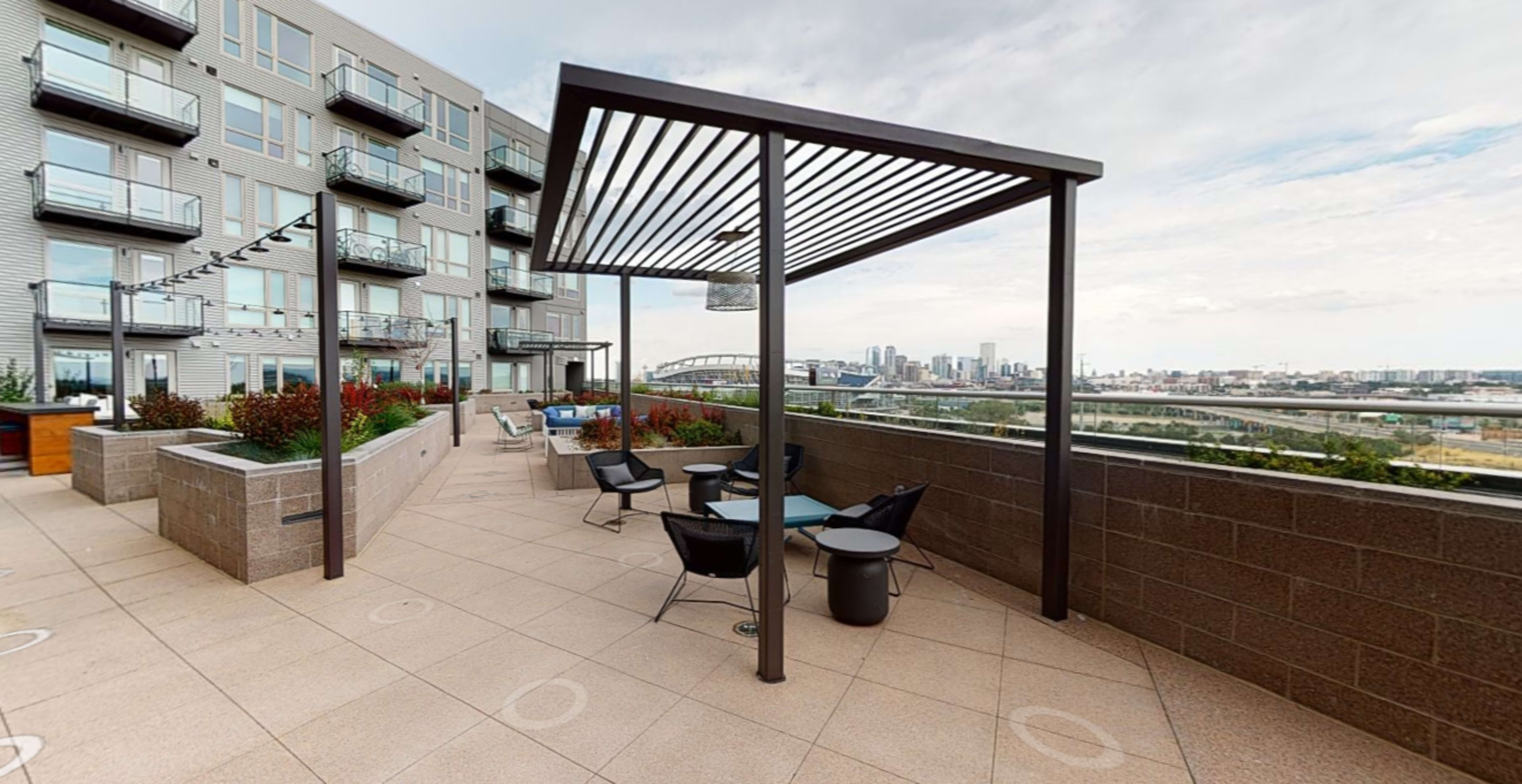 A rooftop terrace with several chairs and tables, surrounded by planters and a view of a city skyline.
