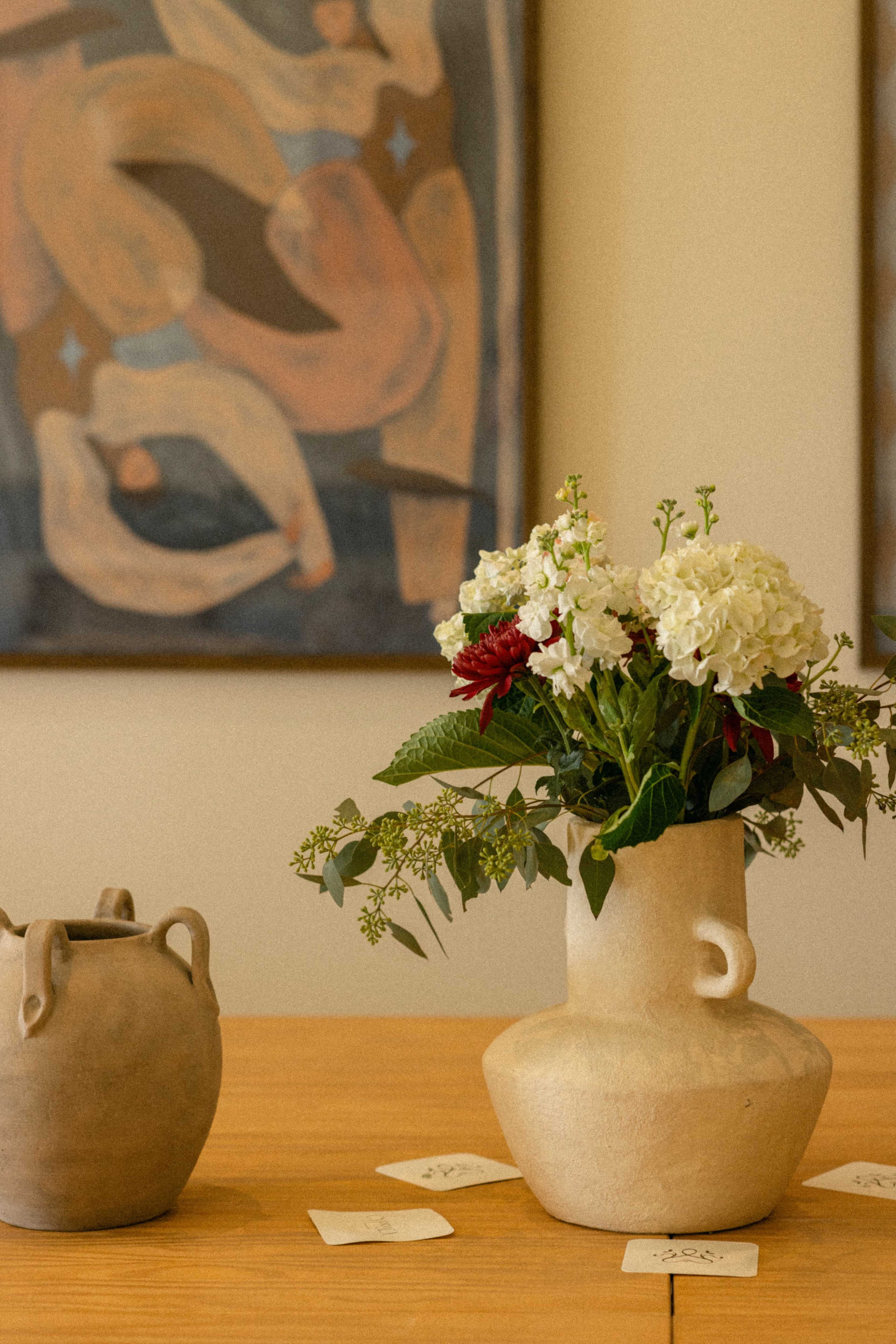 A light-colored vase with flowers sits on a wooden table in front of an abstract painting.