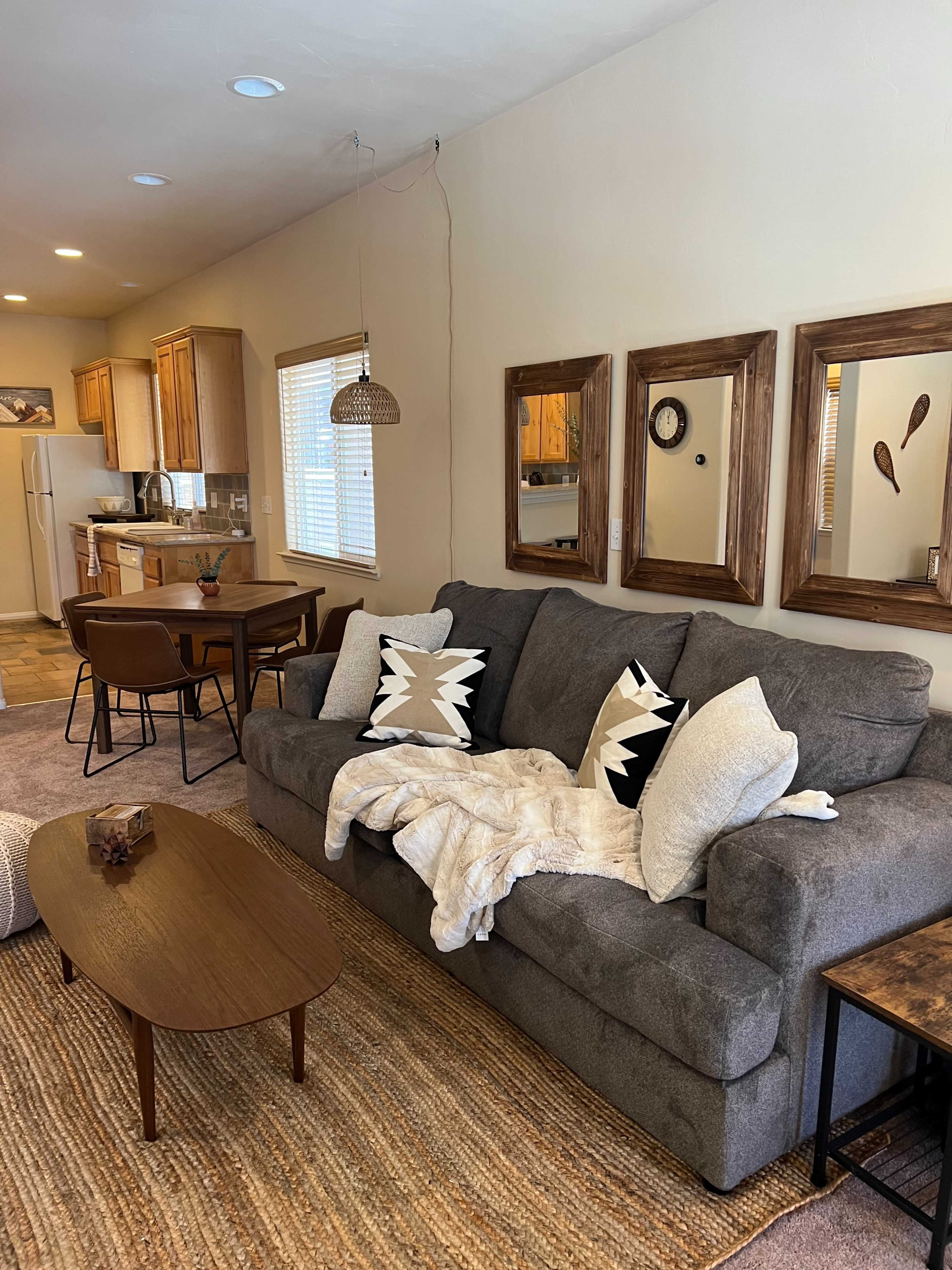 A living room with a gray couch, decorative pillows, a coffee table, and a kitchen area visible in the background.