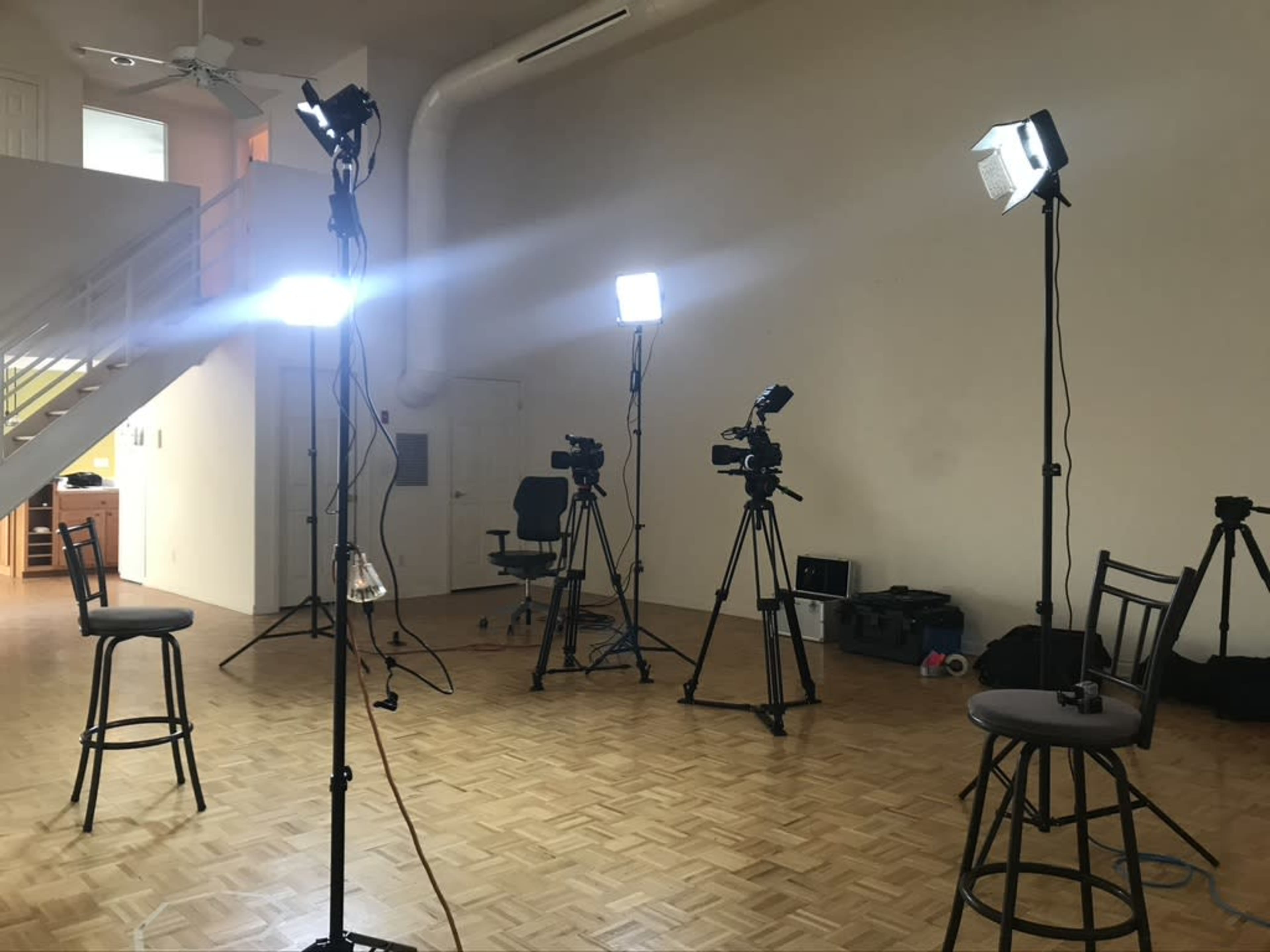 A well-lit recording studio with multiple camera setups and bar stools arranged in a spacious, empty room.