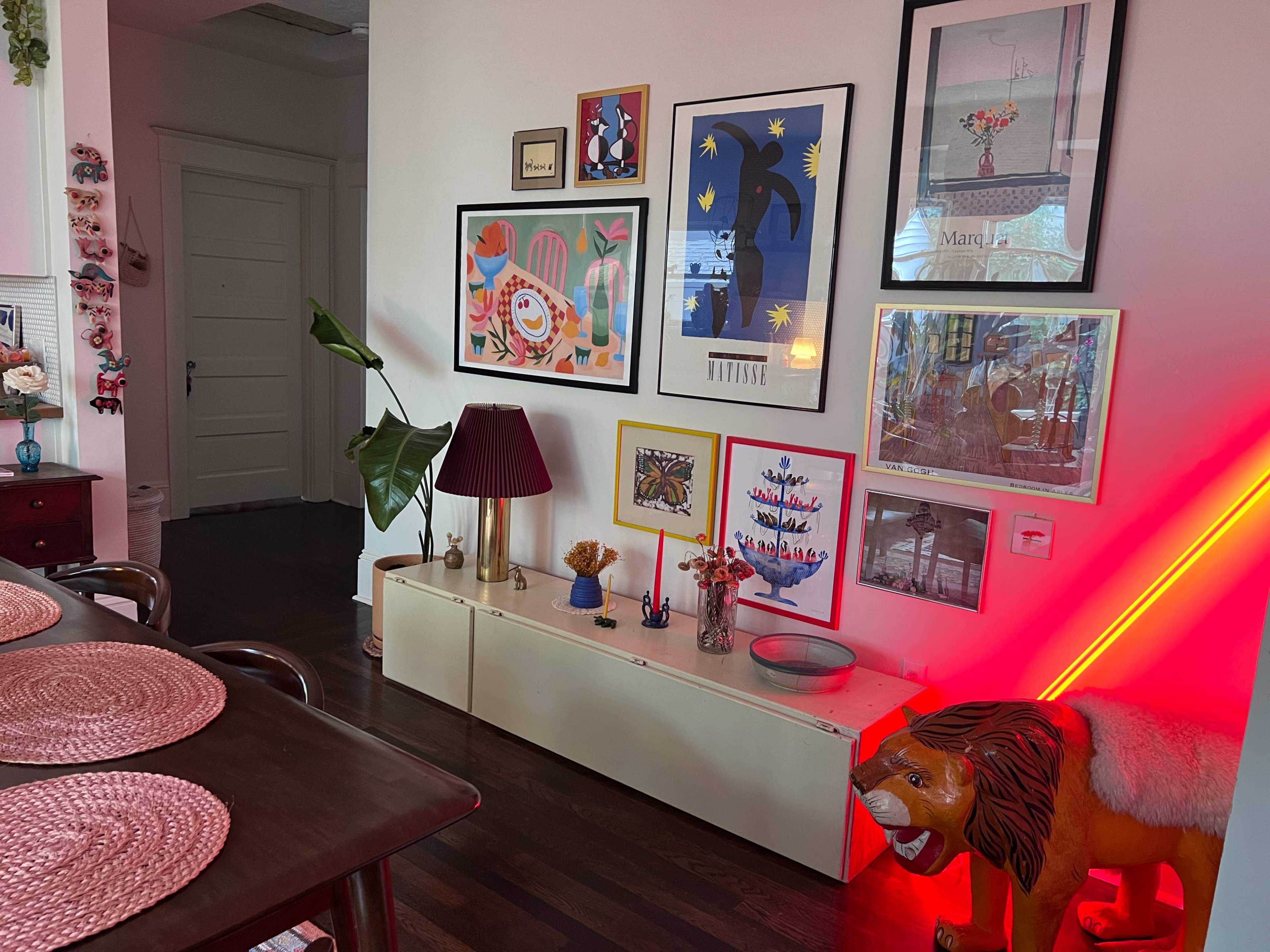 A brightly decorated corner of a room featuring a variety of framed artworks, a potted plant, a lamp, and a colorful lion sculpture on the floor.