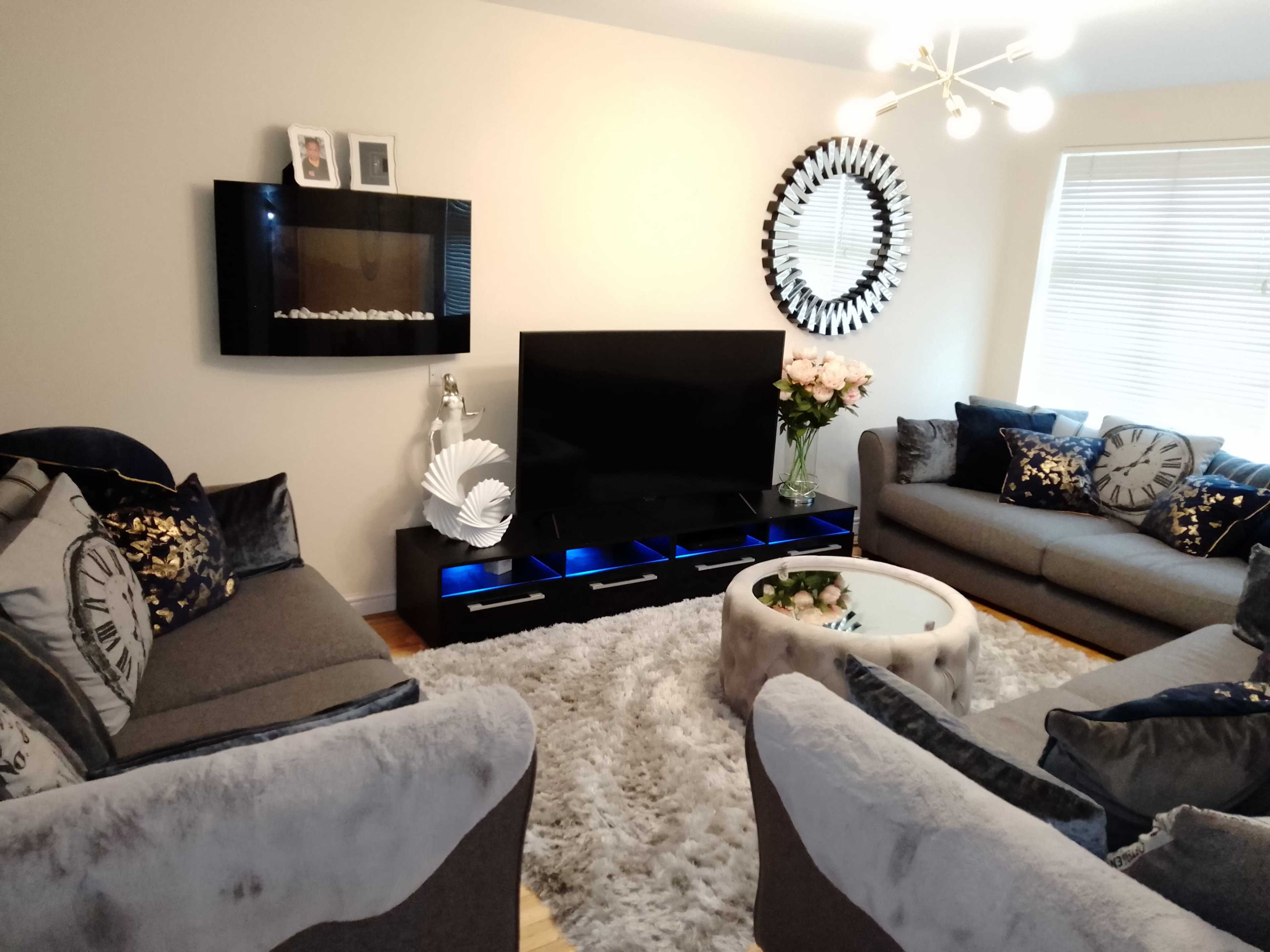 The living room features a modern setup with two gray sofas, a television mounted on the wall, and a round coffee table on a plush rug.