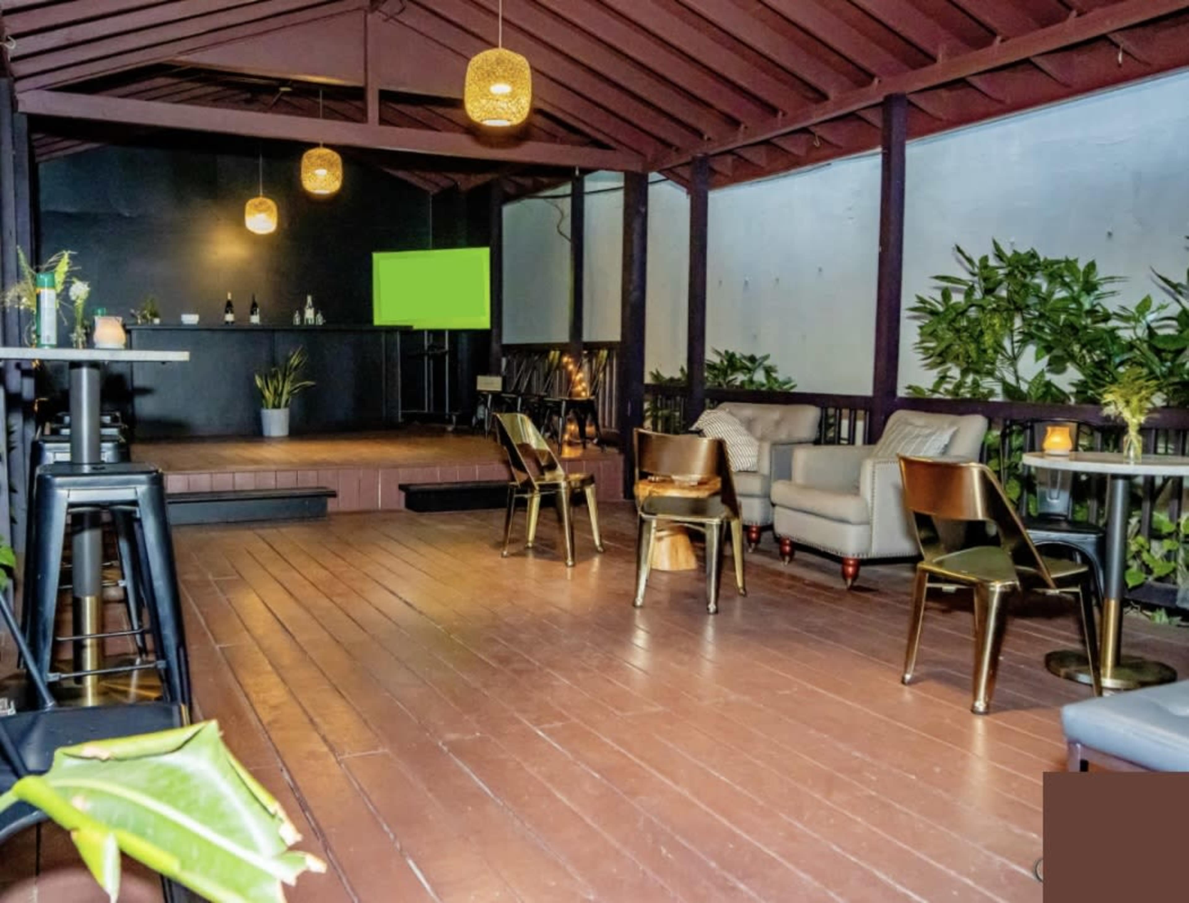 The image shows an indoor seating area with wooden floors, a stage at the back, and a few tables and chairs arranged around the space, accented by hanging light fixtures.
