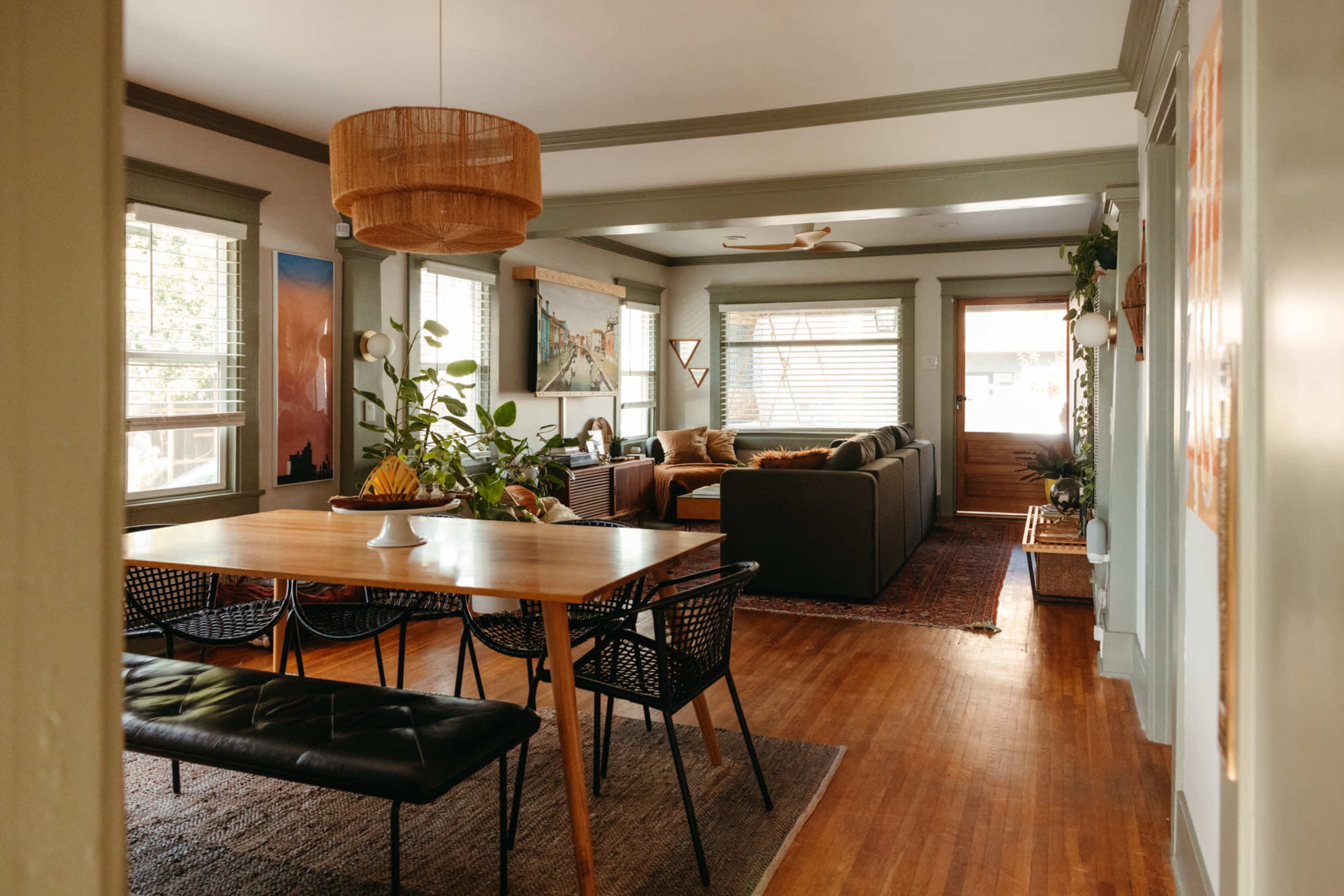 A cozy living and dining area with a wooden dining table, black chairs, and a comfortable sofa arranged around a coffee table, illuminated by natural light from the windows.
