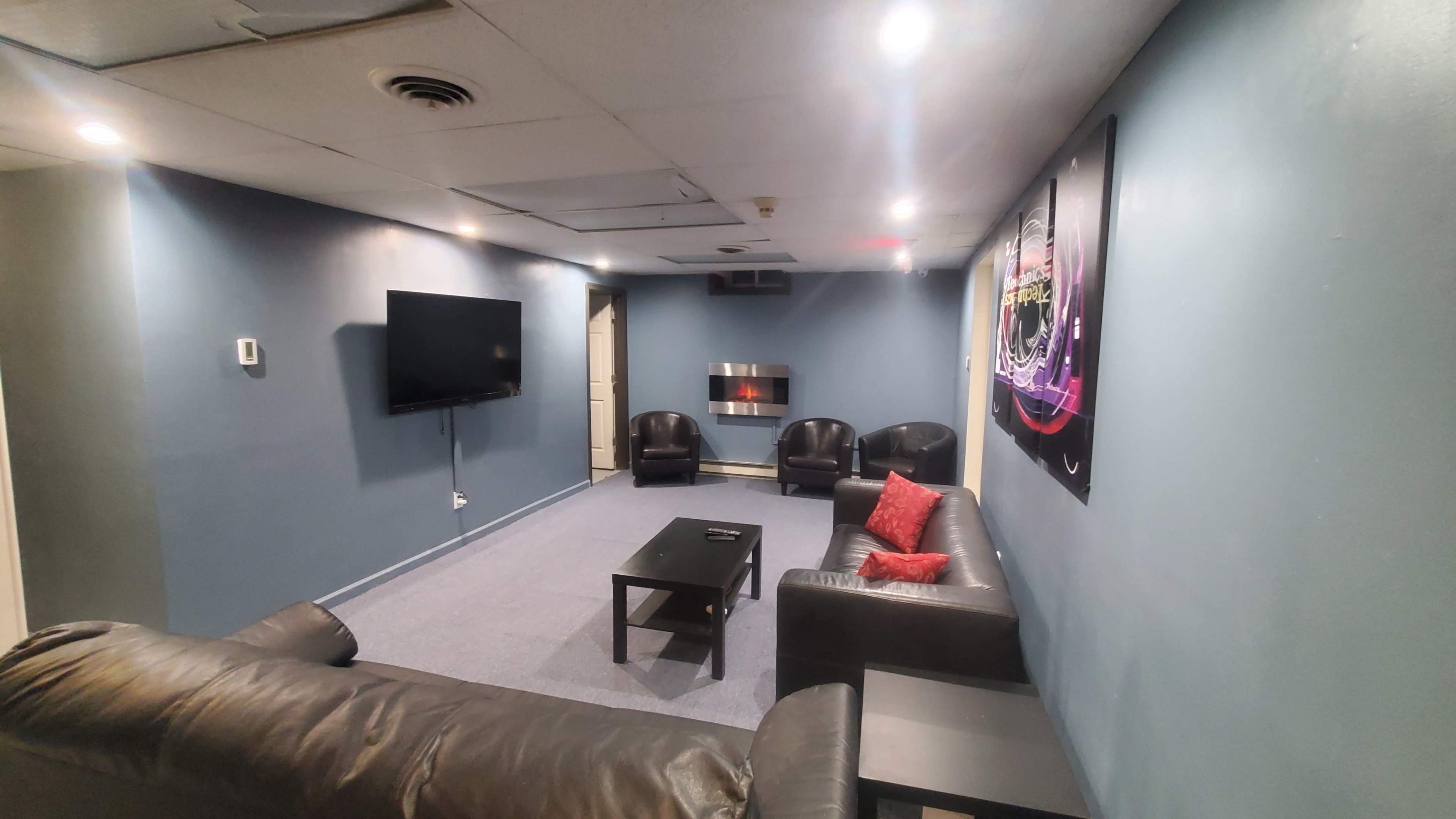 The image shows a lounge area with a television, a fireplace, black leather armchairs, and a couch, all set against a blue wall.