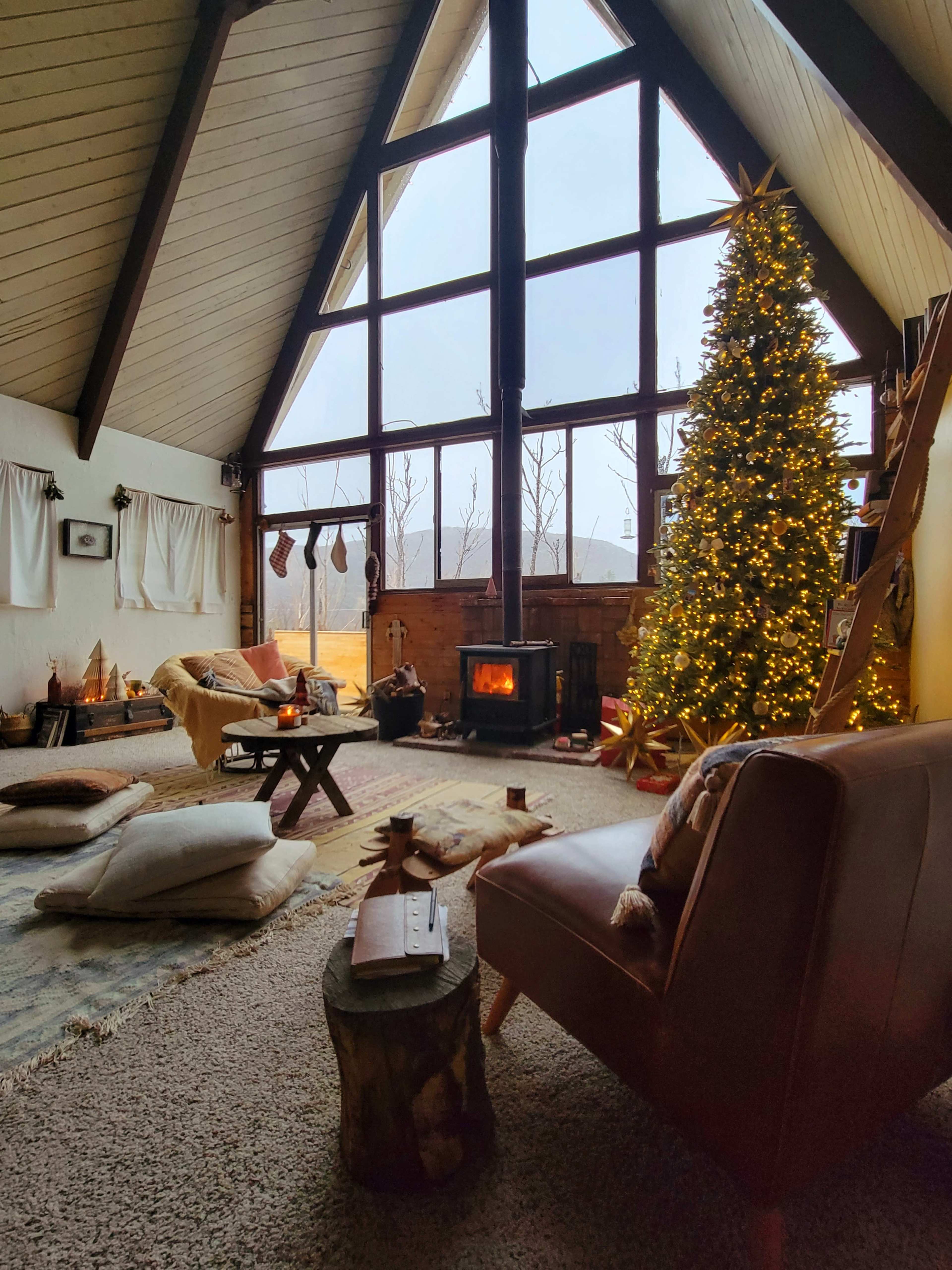 A cozy living room features large windows, a decorated Christmas tree, a fireplace, and comfortable seating.