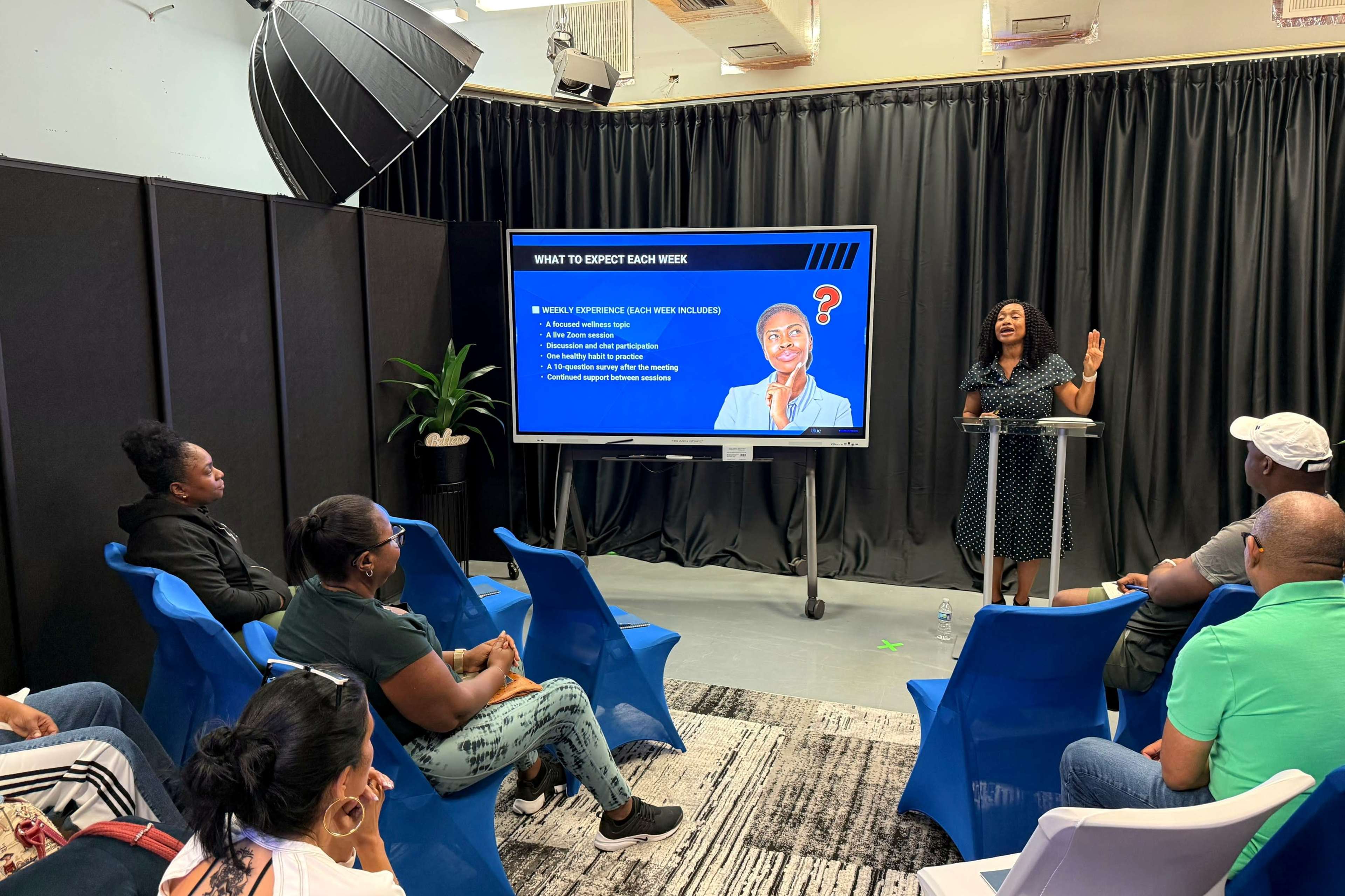 A woman stands in front of a screen presenting to an audience seated in chairs, discussing weekly expectations for a program or event.