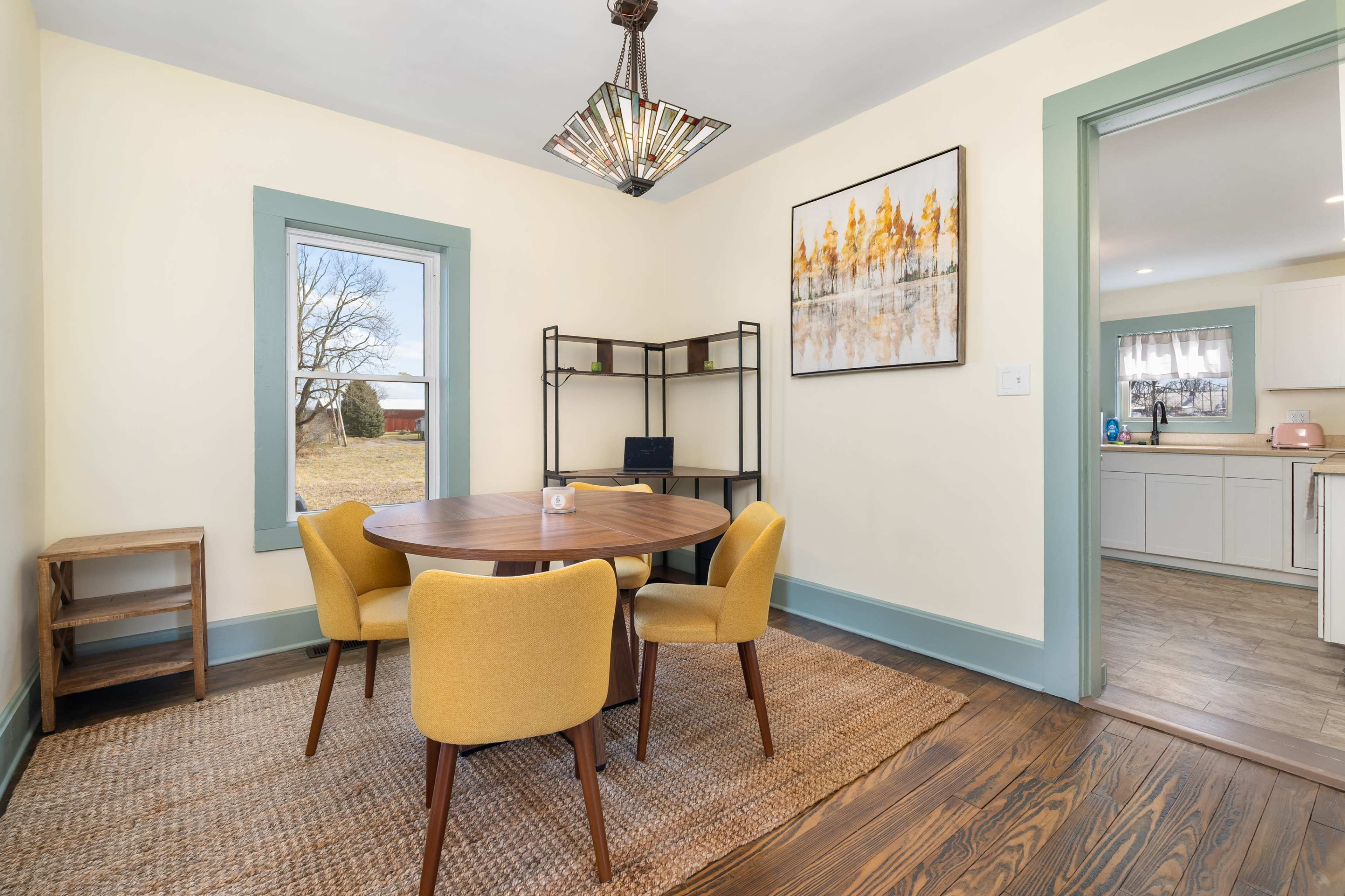 The image shows a dining area with a round table surrounded by yellow chairs, a bookshelf, and a window looking out to a field.