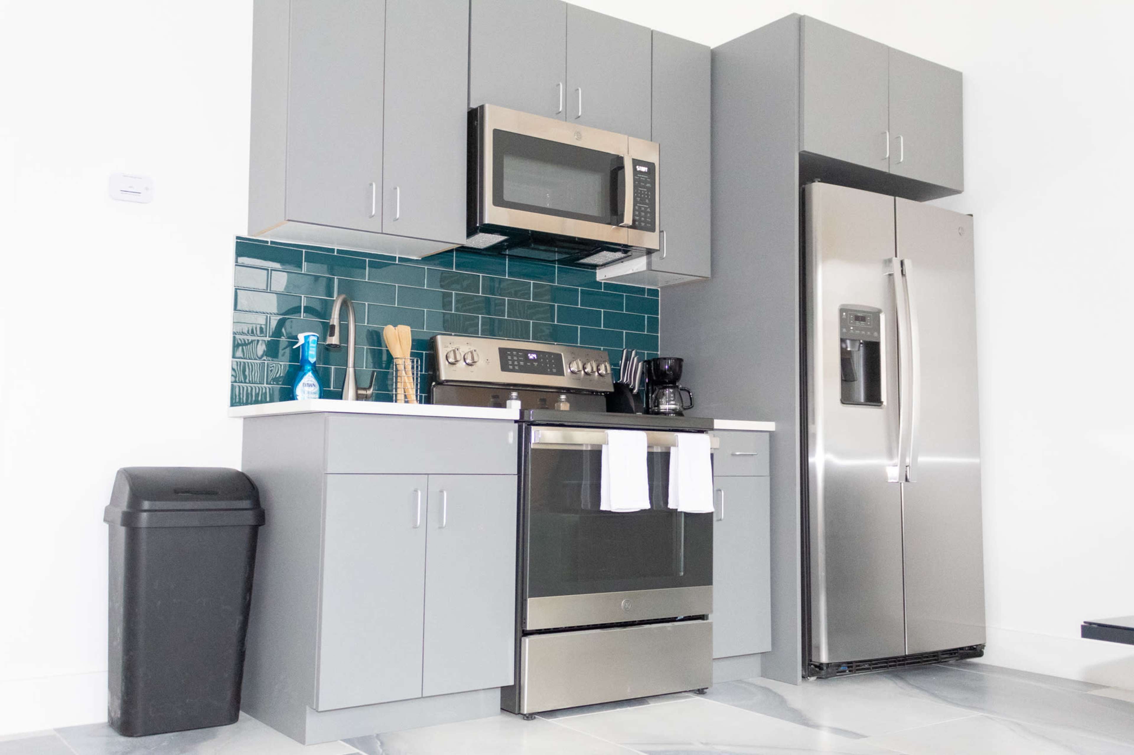 The image shows a modern kitchen with gray cabinets, stainless steel appliances, and a teal tiled backsplash.