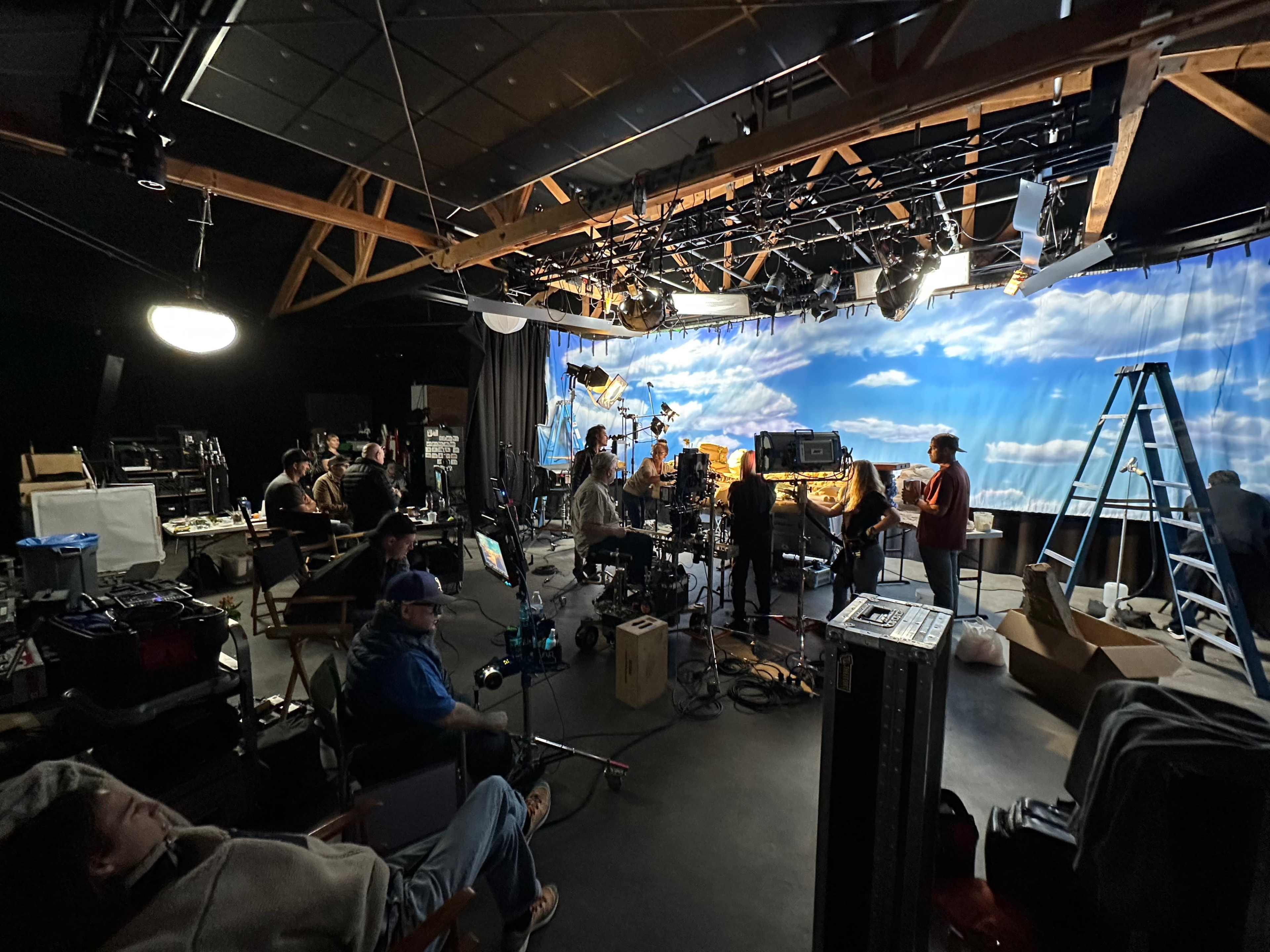 A film crew sets up in a studio with a backdrop of blue skies and clouds, surrounded by equipment and crew members preparing for a shoot.