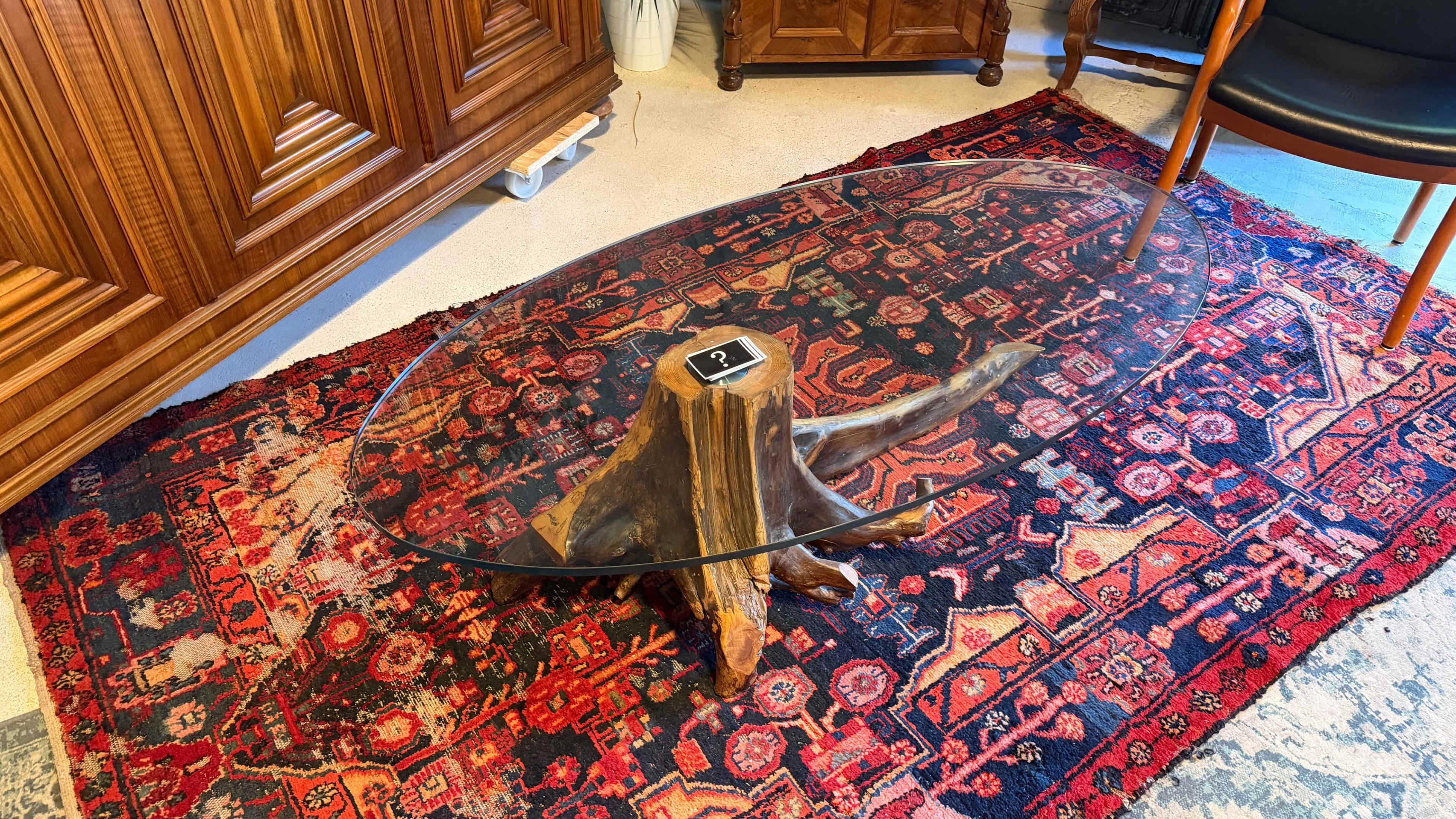 The image features a glass-topped coffee table supported by a wooden stump, placed on a colorful patterned rug in an indoor setting.