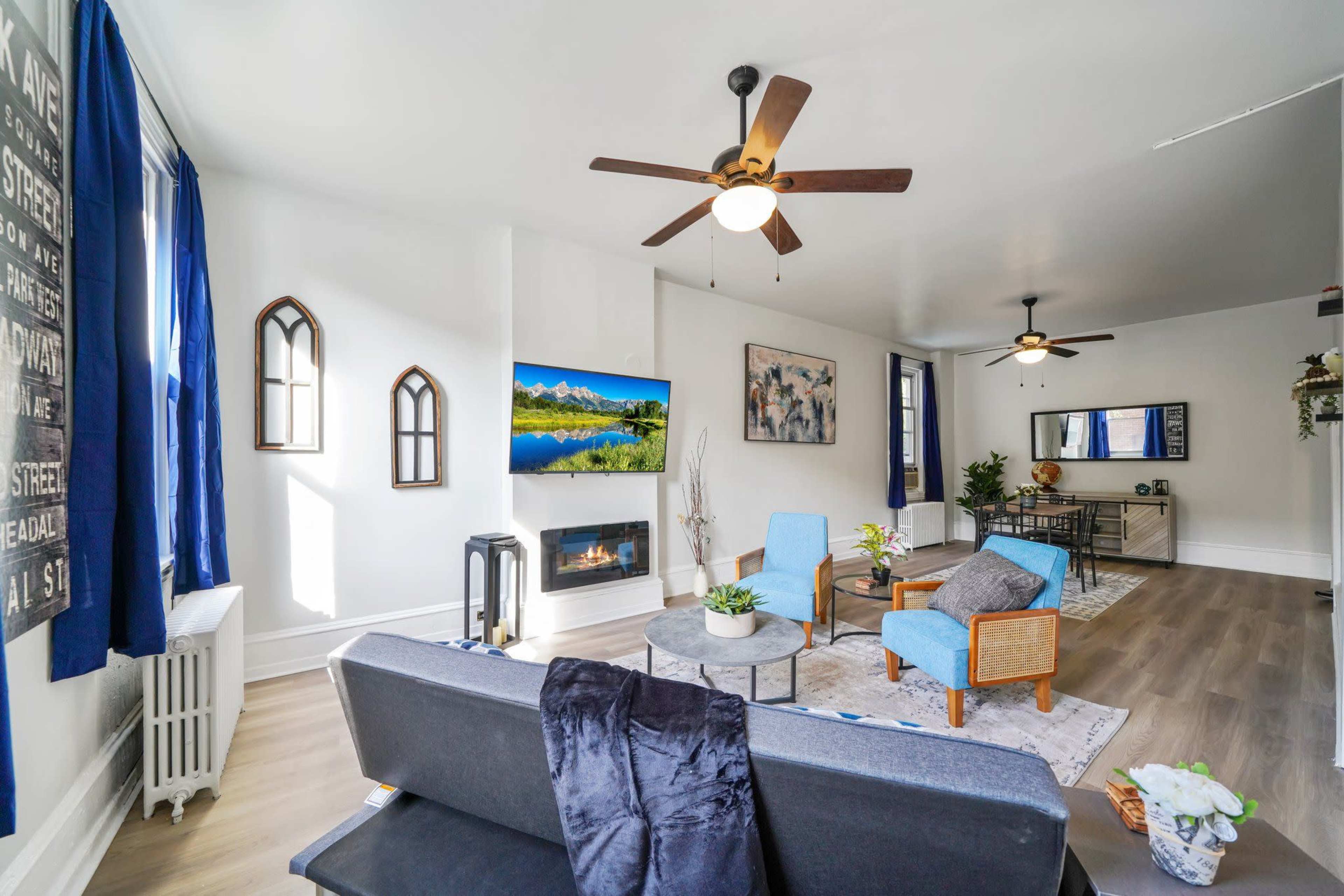A modern living room features a gray sofa, two blue chairs, a television mounted on the wall, and a decorative fireplace.