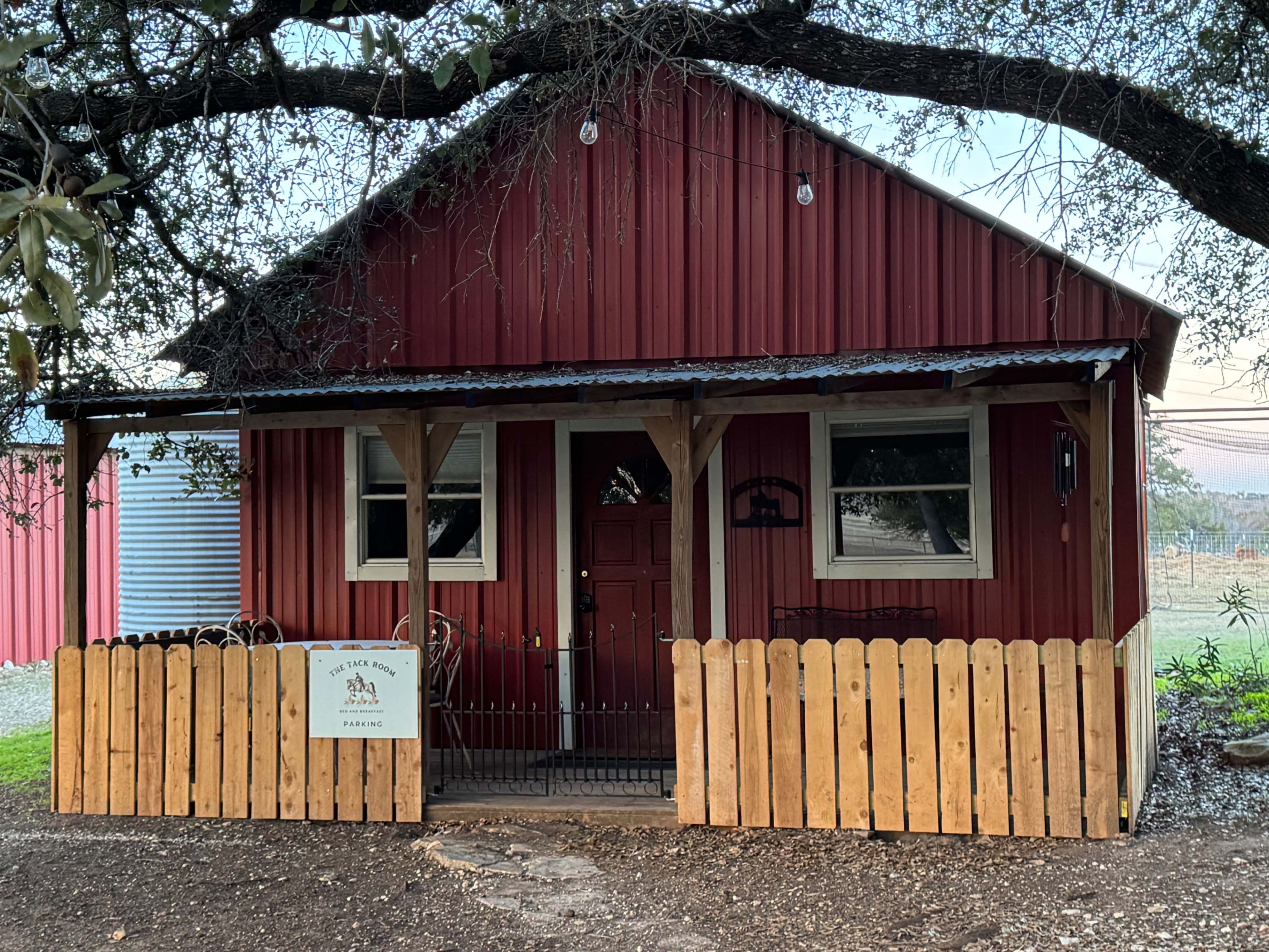 The image shows a small red barn-style cabin with a wooden fence in front, surrounded by grass and shaded by a large tree.