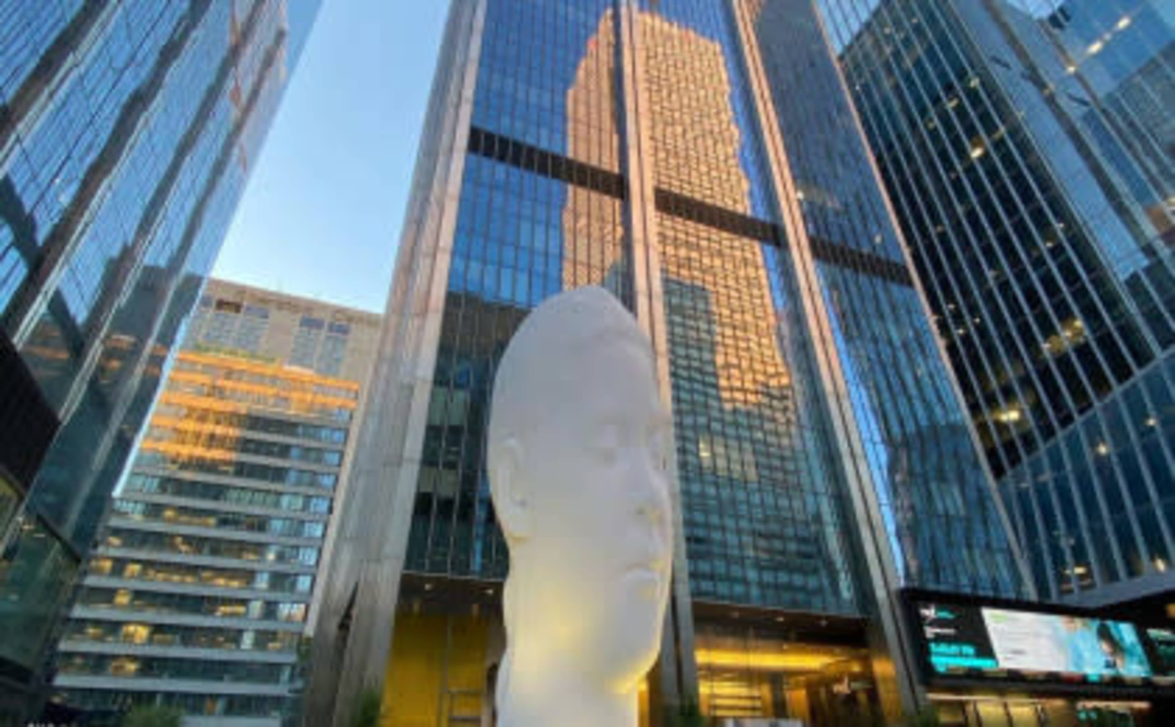 A large white sculpture of a head stands in front of reflective glass skyscrapers.