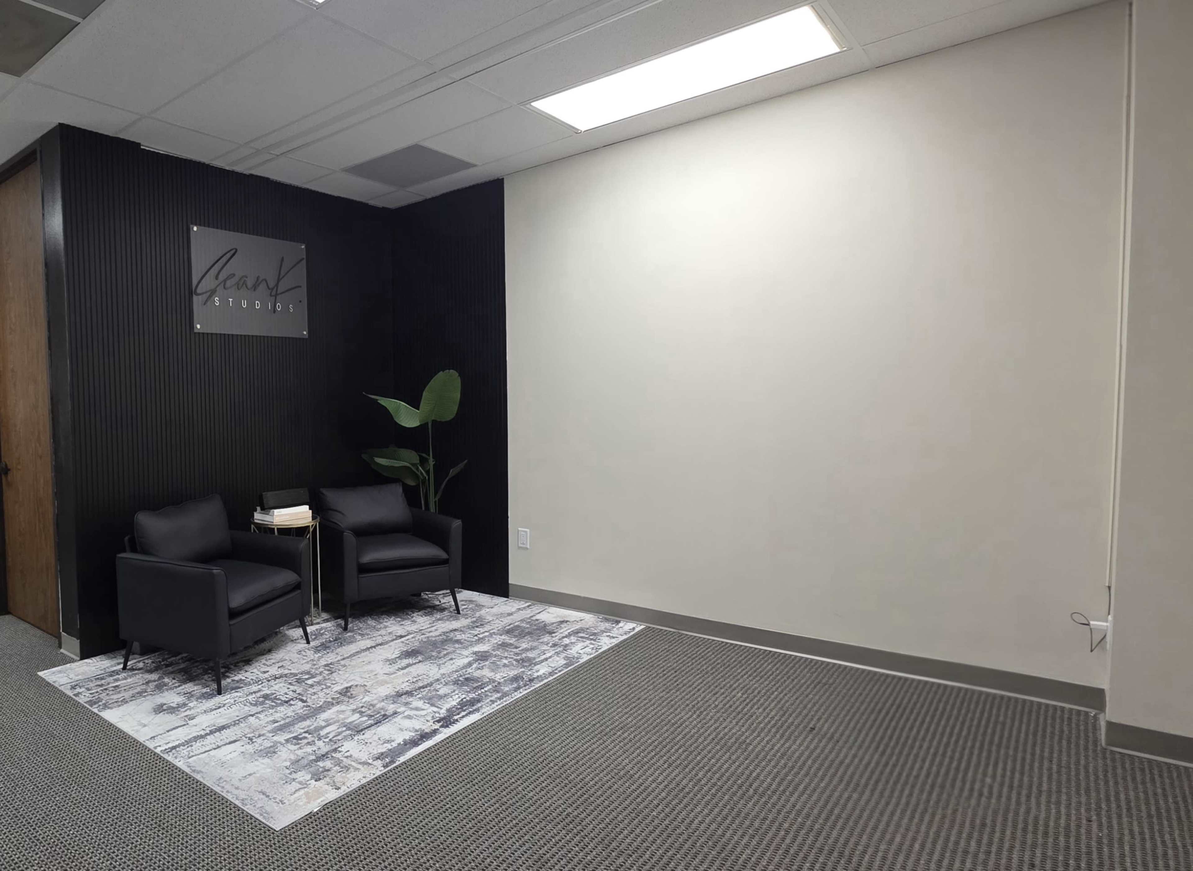 The image shows a waiting area with two black chairs, a small side table, and a decorative rug against a plain, well-lit wall with a sign that says "Leah's Studio."