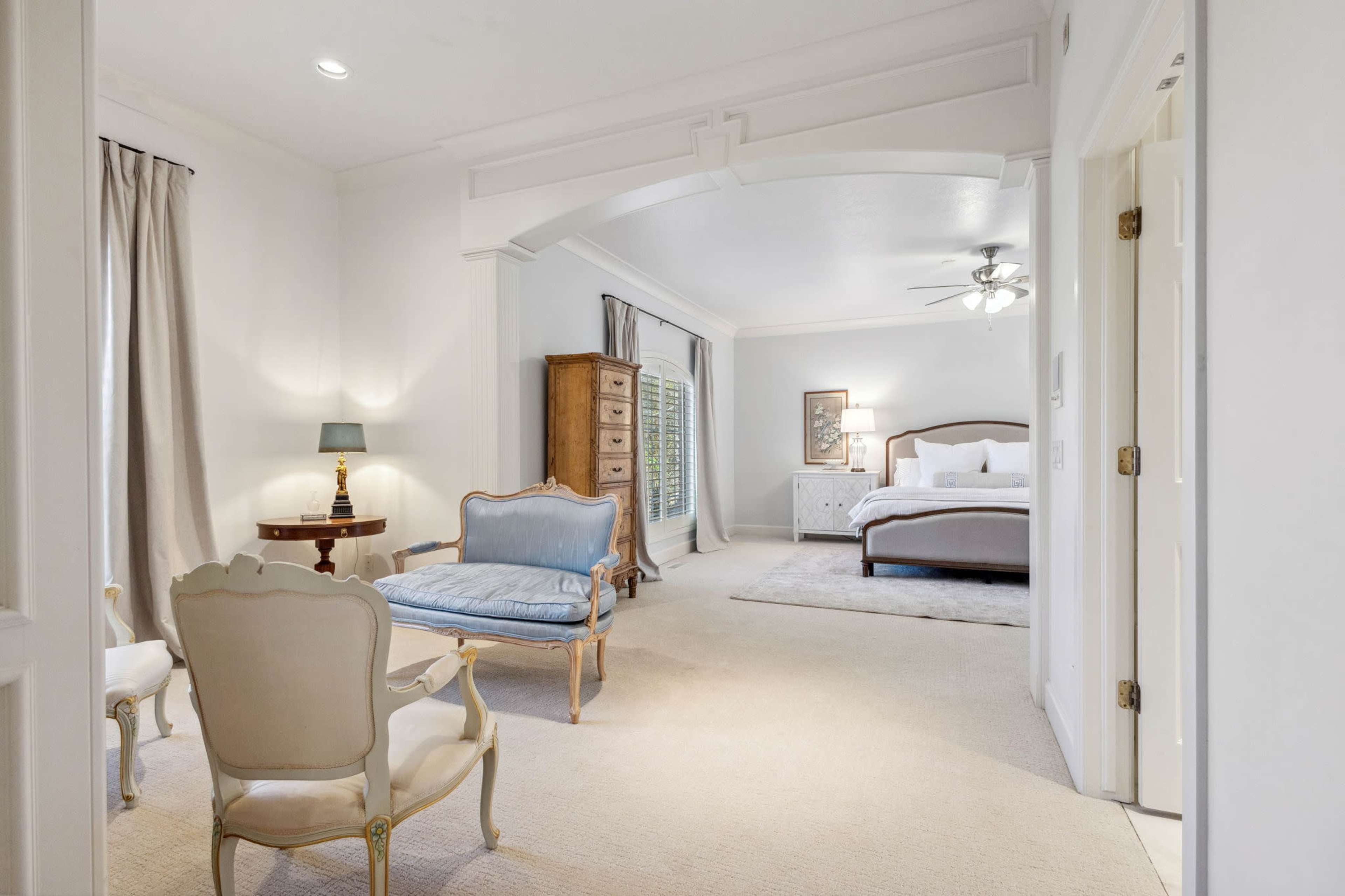 A spacious room features a bed in the corner, with a seating area that includes a blue settee and a white armchair, all under soft lighting.