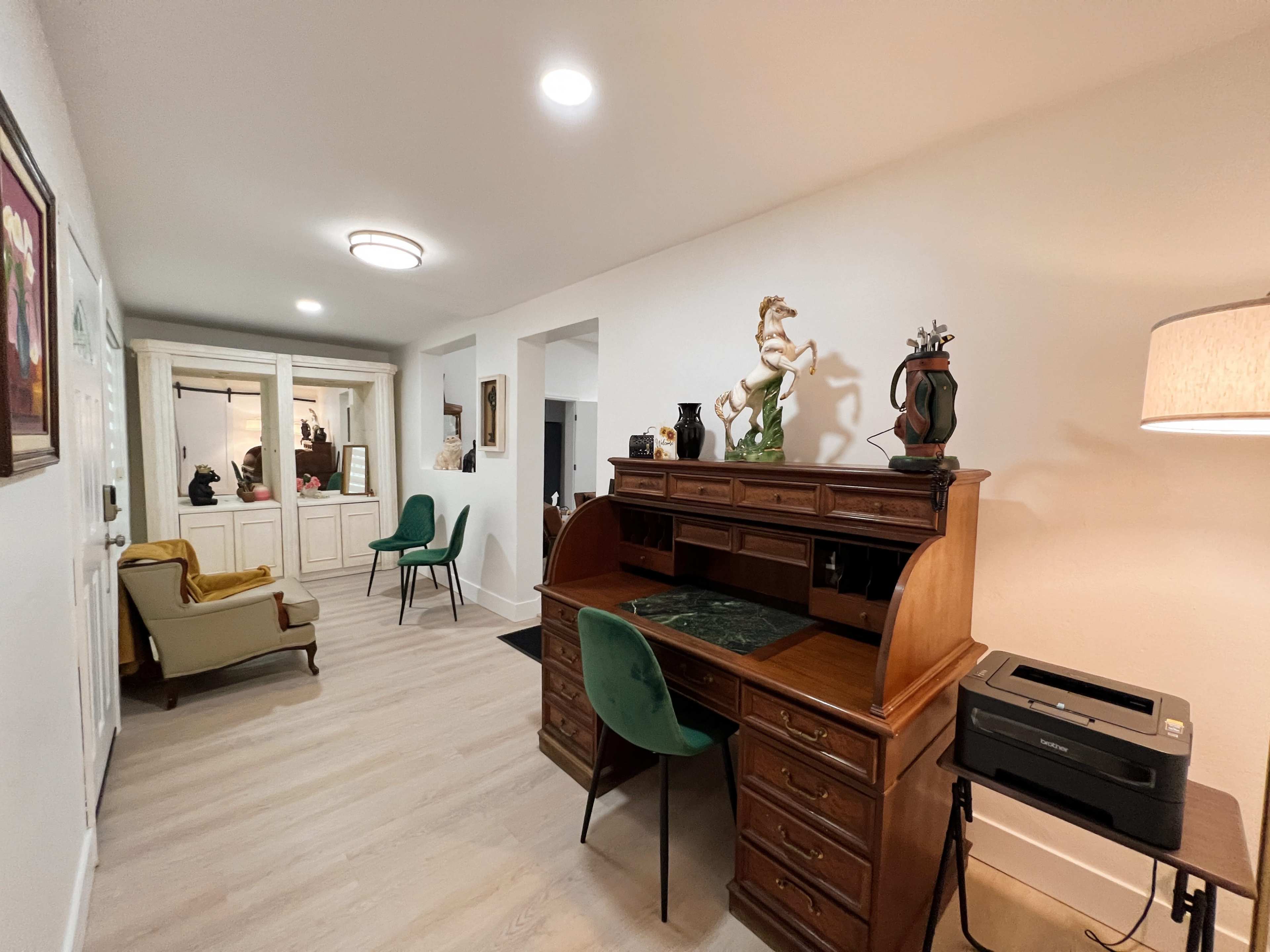 A hallway featuring a wooden desk with a green chair, a decorative lamp, and various artwork on the walls.