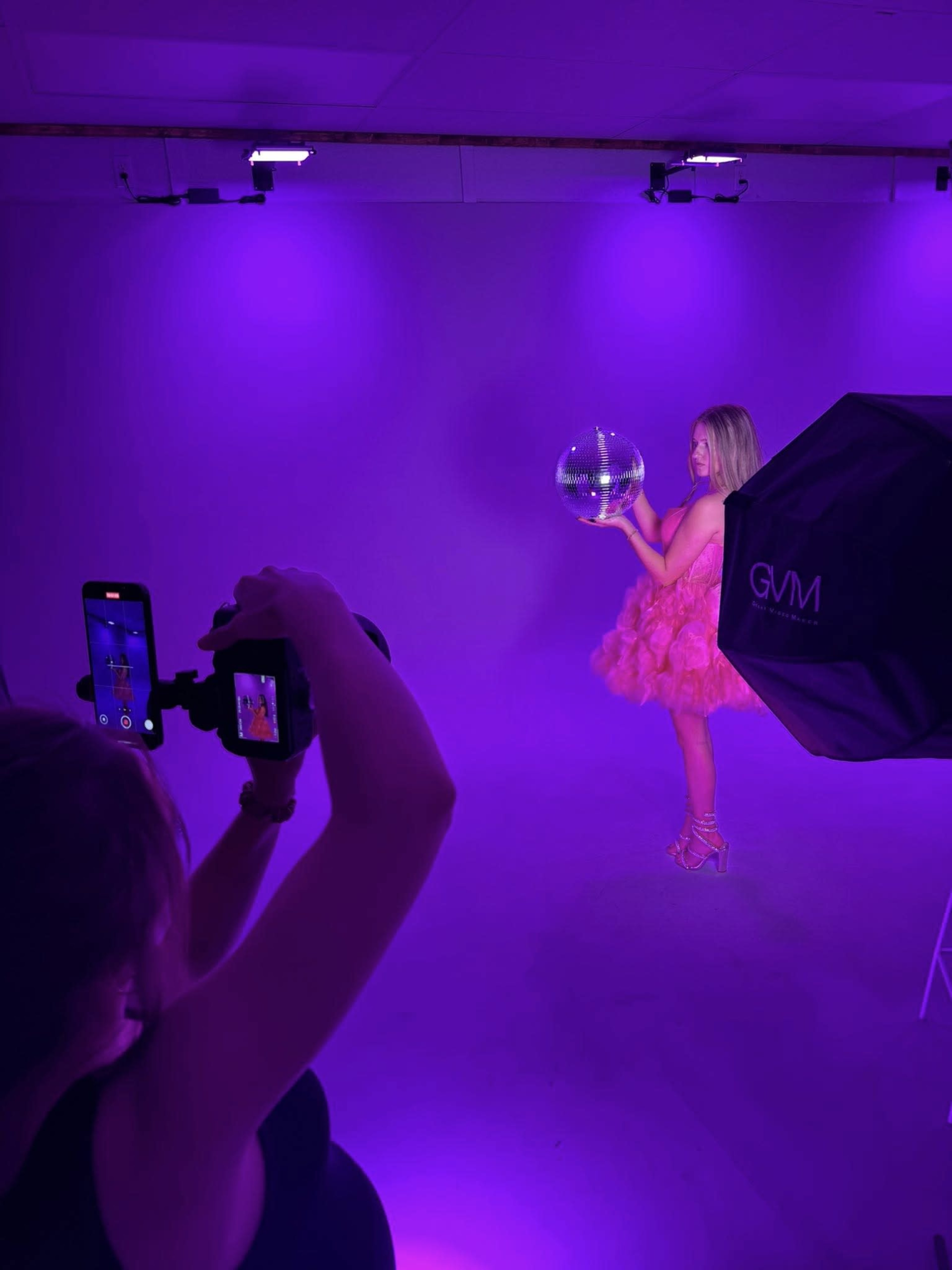 A person in a pink, fluffy dress holds a reflective disco ball while another individual photographs them against a purple-lit backdrop.