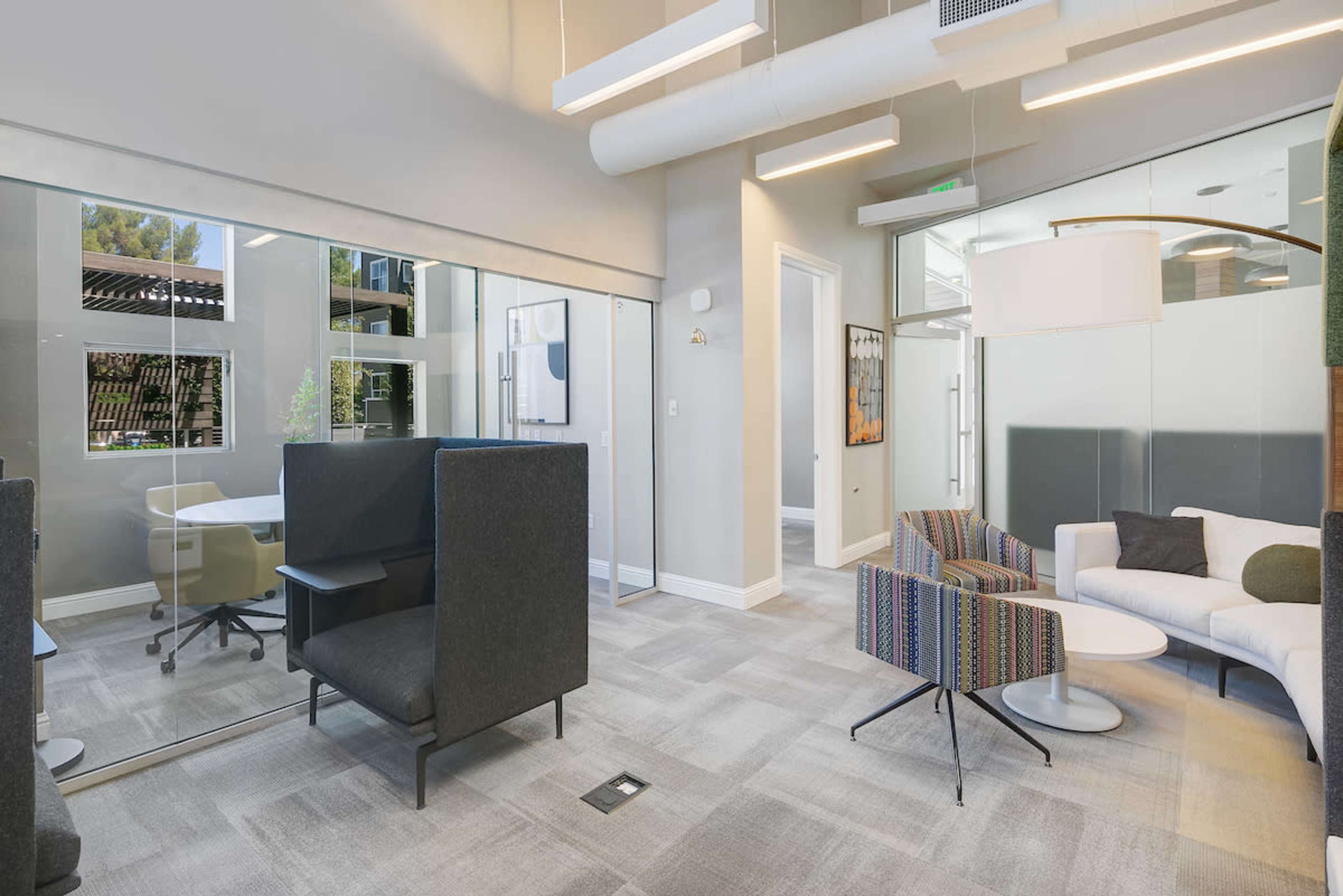The image shows a modern office space featuring a seating area with a couch and chairs, glass partitions, and a meeting room.