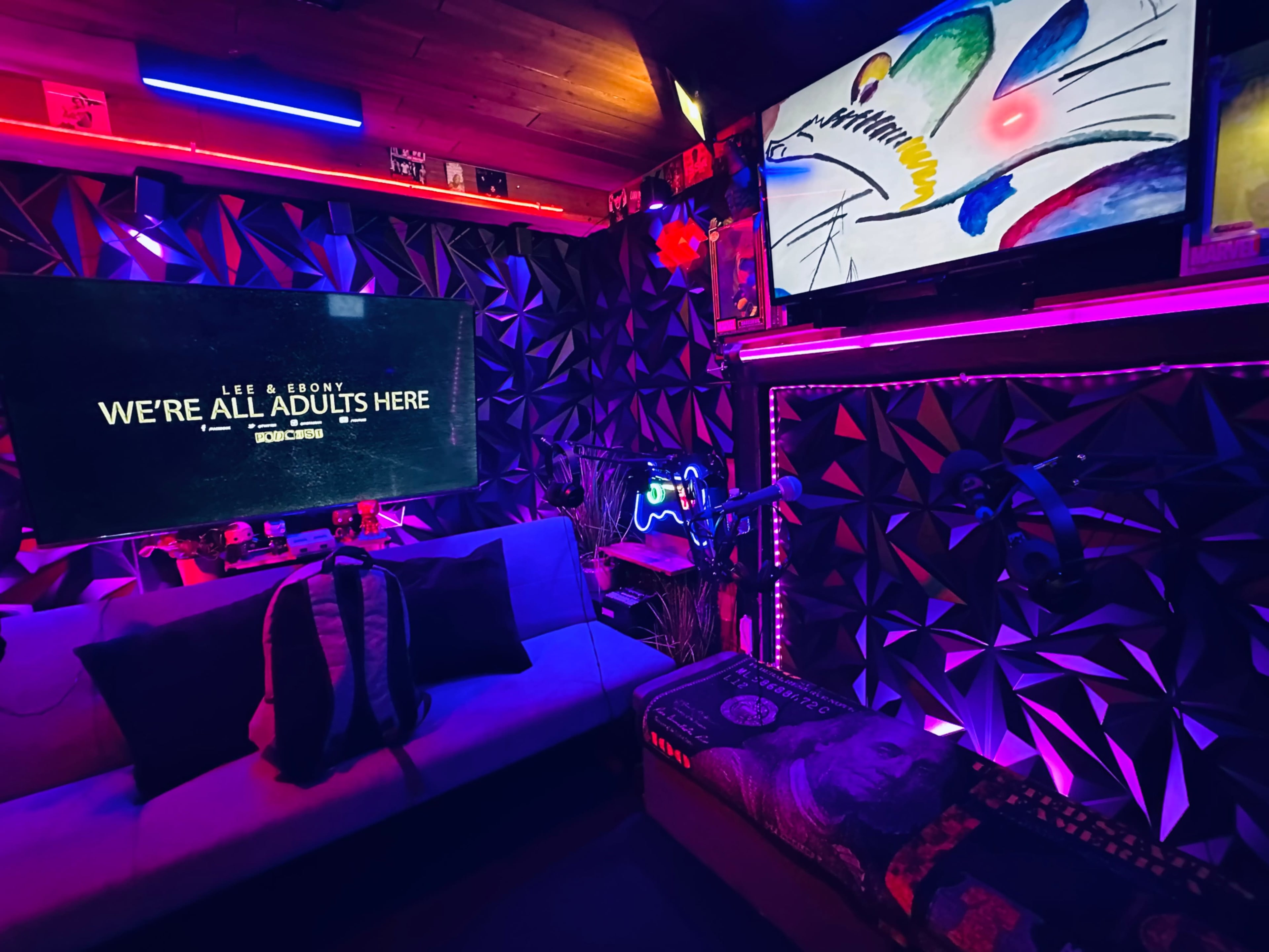 The image shows a dimly lit room with colorful LED strip lighting, a large television displaying the text "WE'RE ALL ADULTS HERE," and various decorative elements including artwork and gaming accessories.