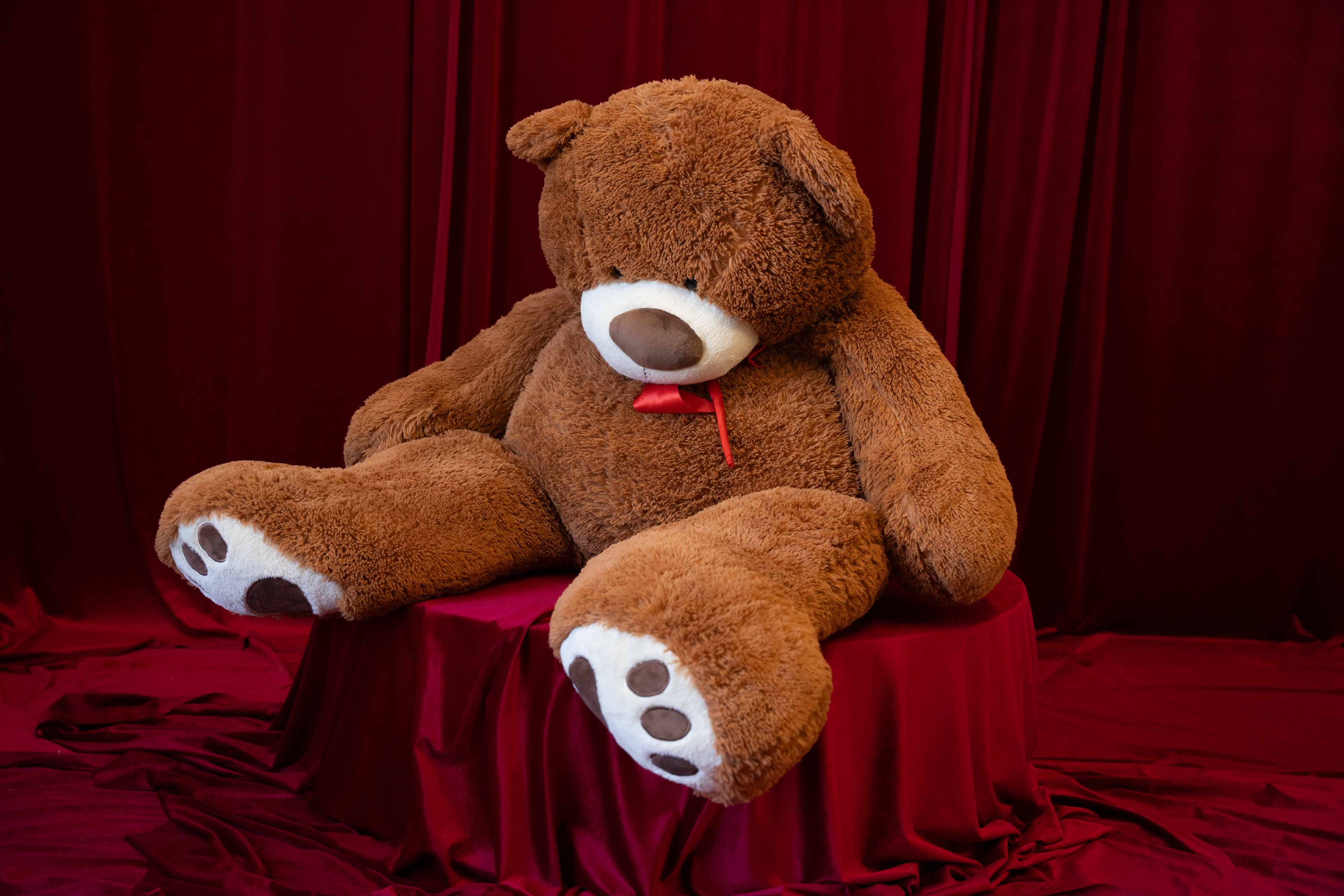 A large, brown stuffed bear is seated on a red pedestal against a backdrop of red fabric.